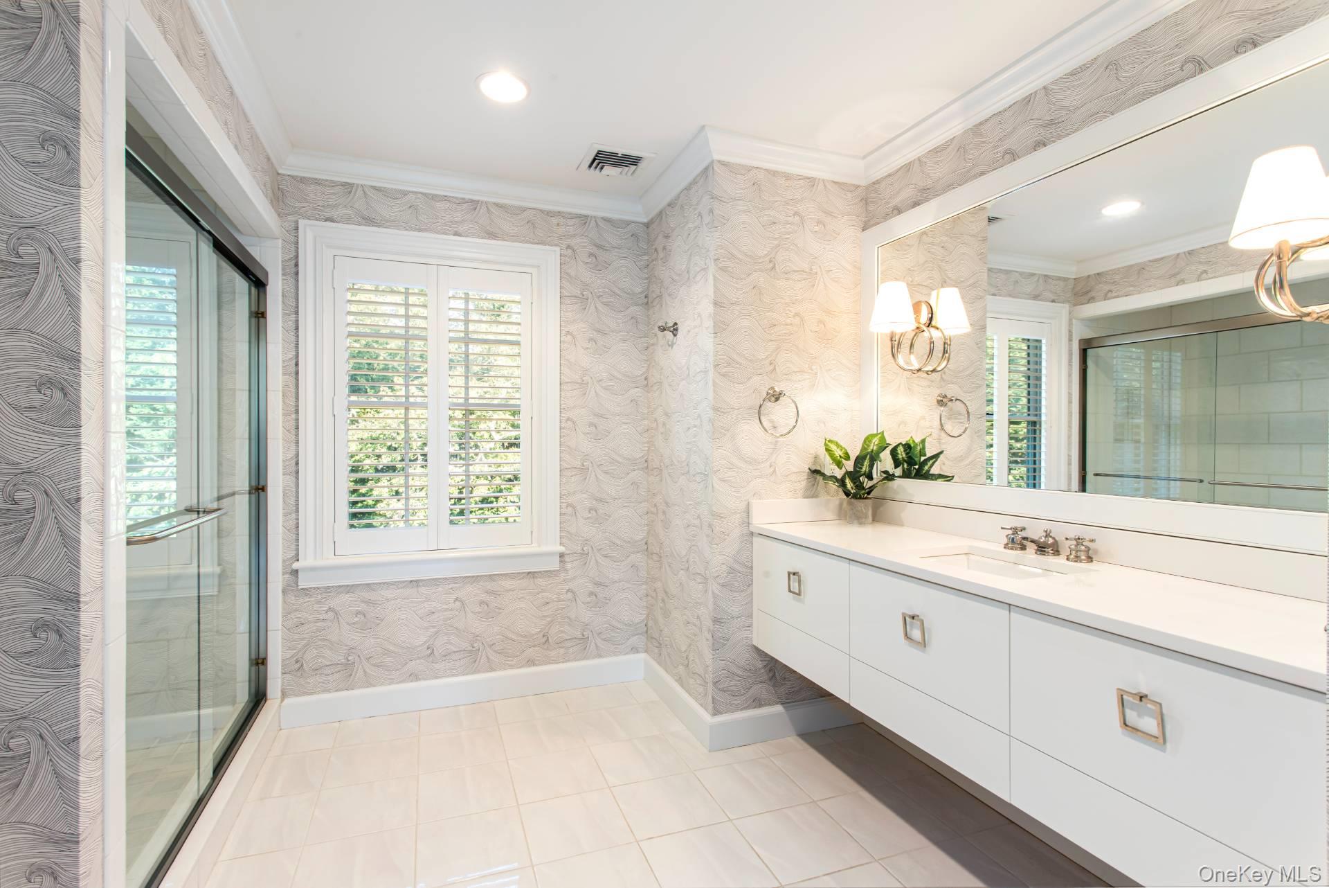 Bathroom with glass shower, long white vanity, shuttered window, and gray patterned wallpaper.