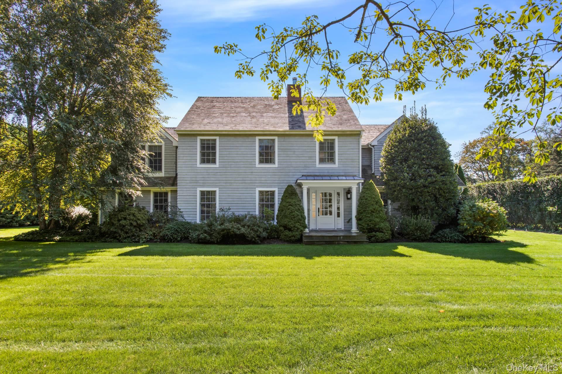 Gray two-story shingle house with a covered front entrance, framed by mature trees and a broad lawn.