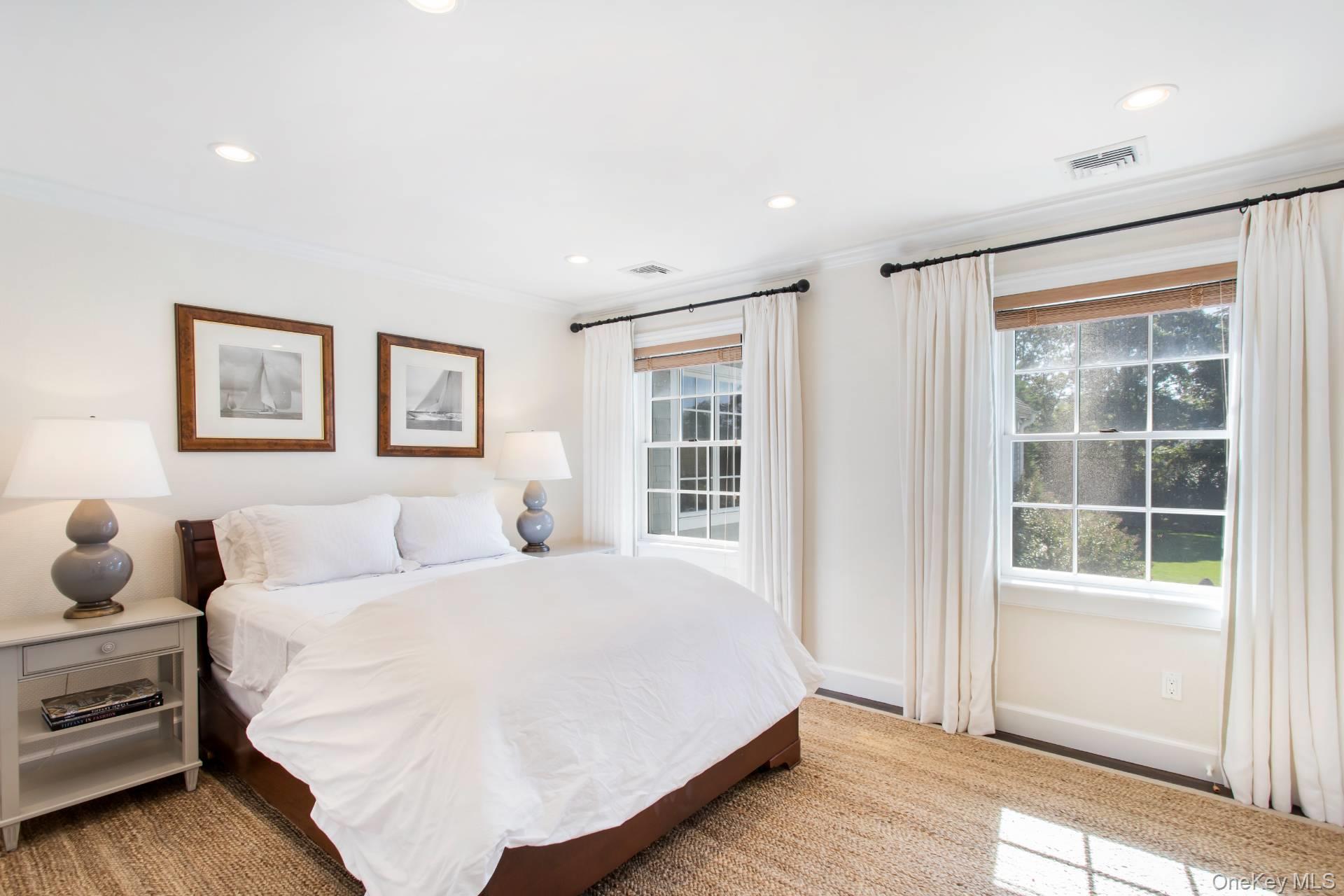 Bright bedroom with a white bed, framed sailboat prints, and windows overlooking greenery.