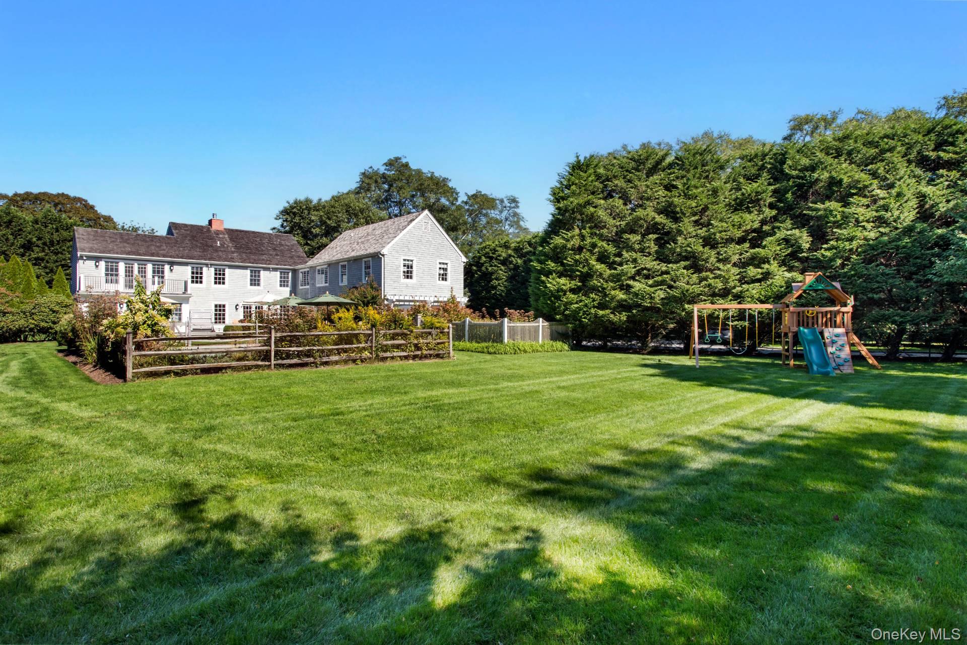 Gray two-story house beyond a broad lawn, with a fenced garden and a wooden playground.