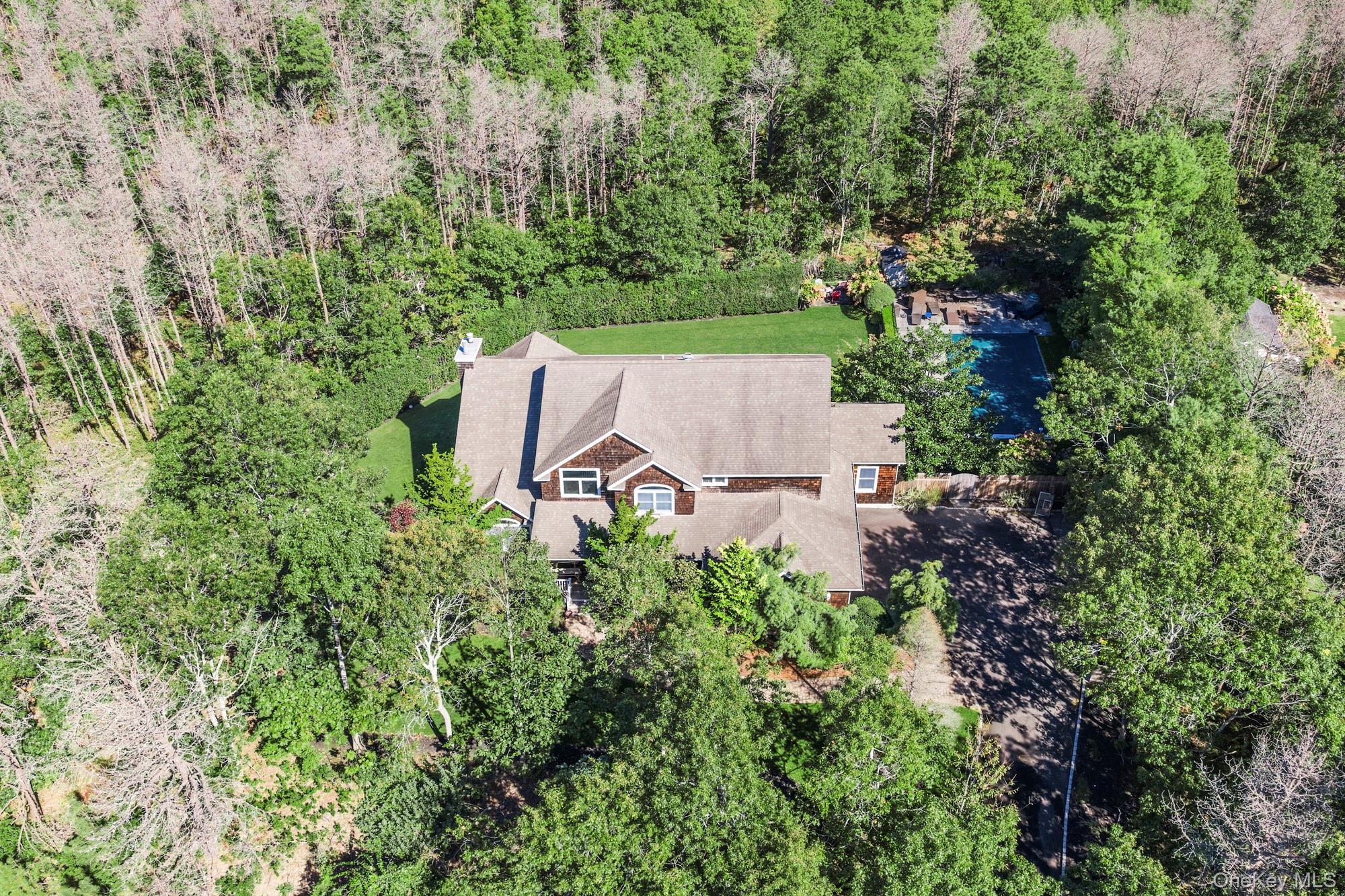 Aerial view of a shingle-sided home amid mature trees, with a backyard pool, patio, and lawn.