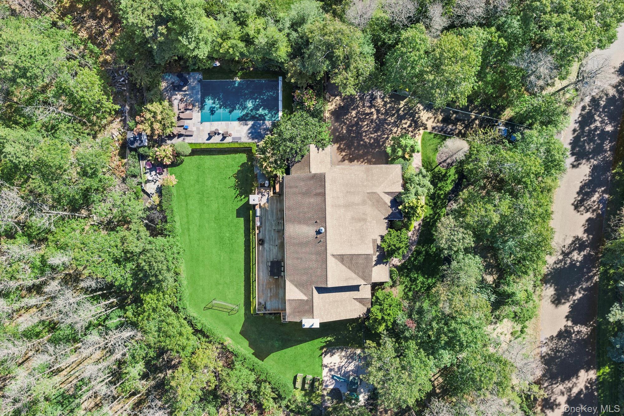 Aerial view of a tree-surrounded home with a green lawn, swimming pool, and paved patio.
