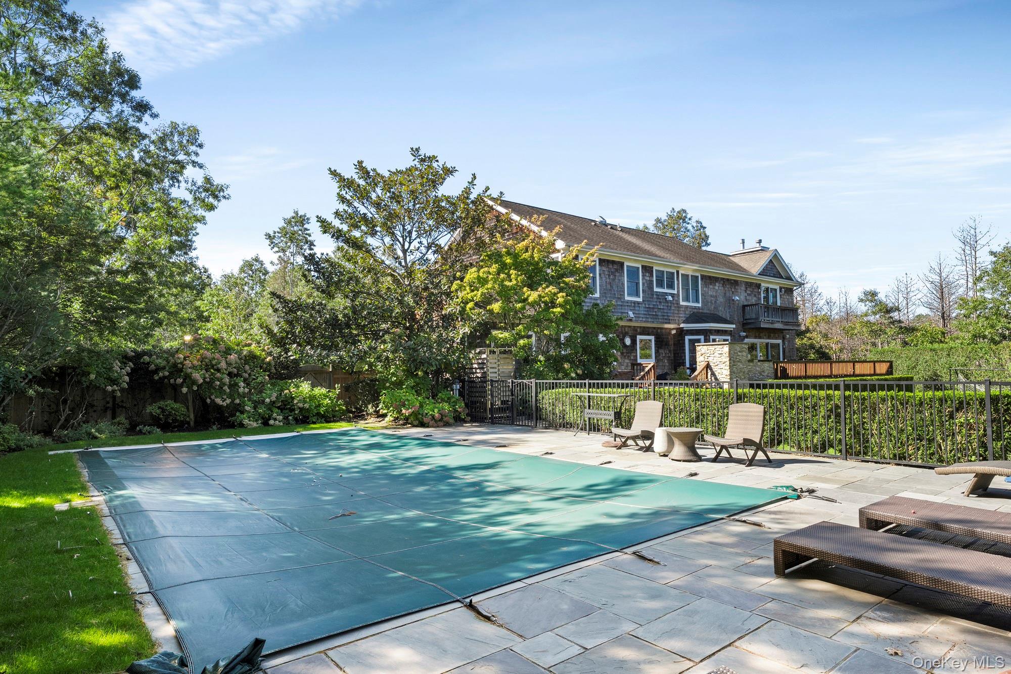 Covered swimming pool beside a shingled house, with patio furniture and mature landscaping.