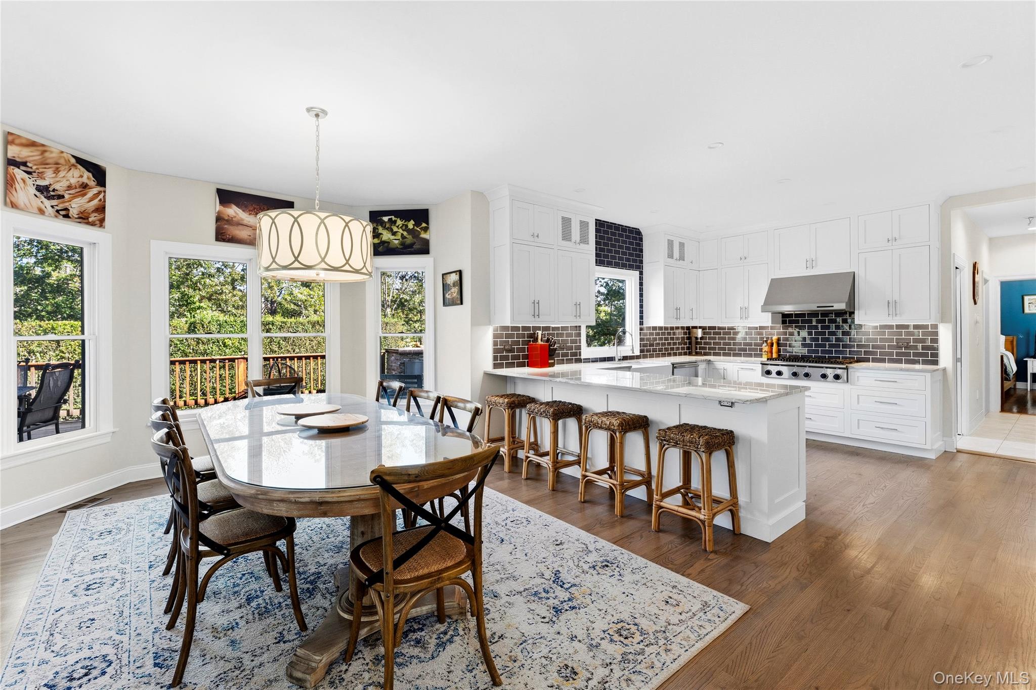 Open kitchen and dining area with white cabinets, dark tile backsplash, island stools, and windows overlooking trees.