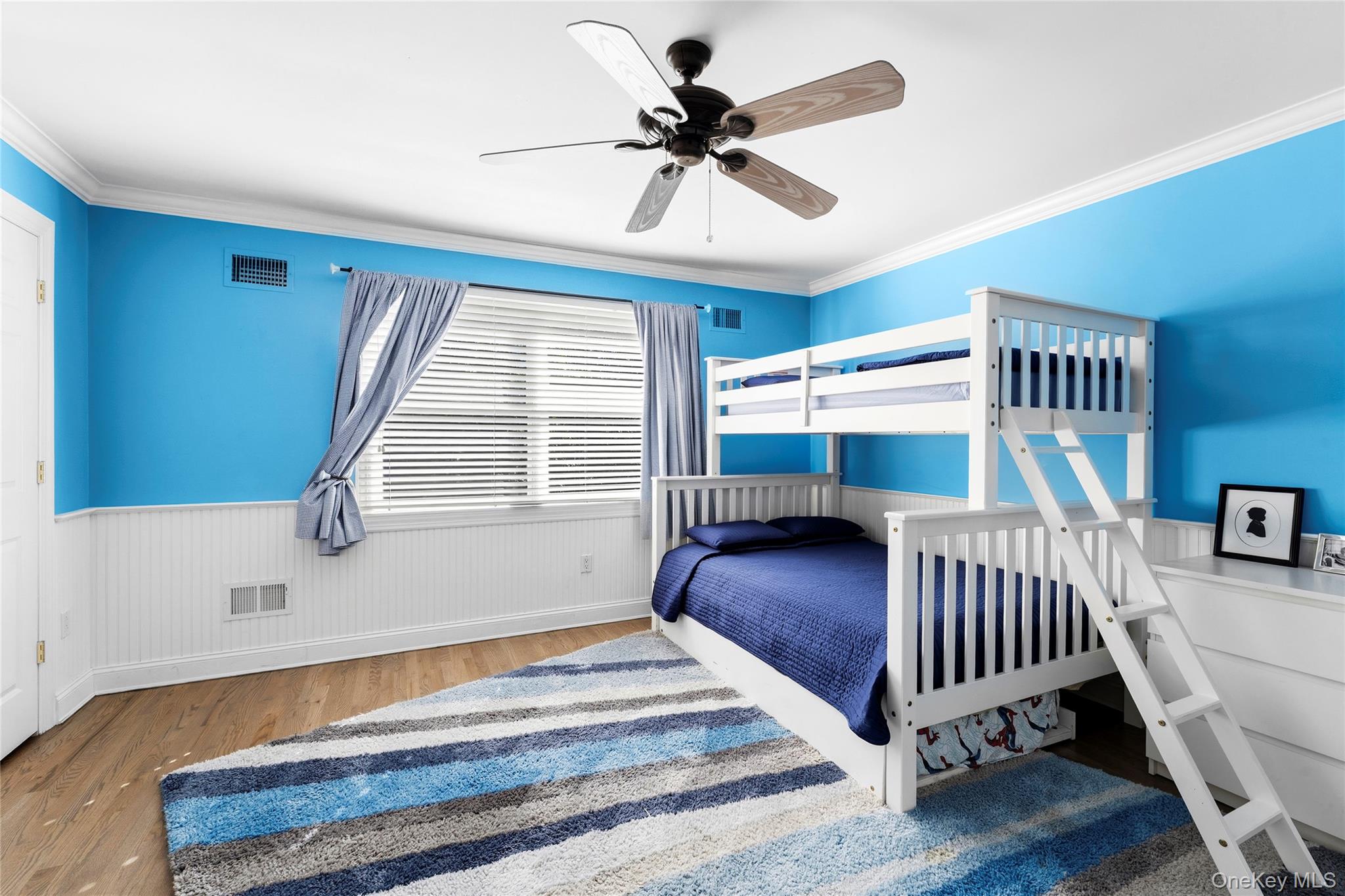 Blue bedroom with a white loft bunk bed, striped rug, and curtained window.