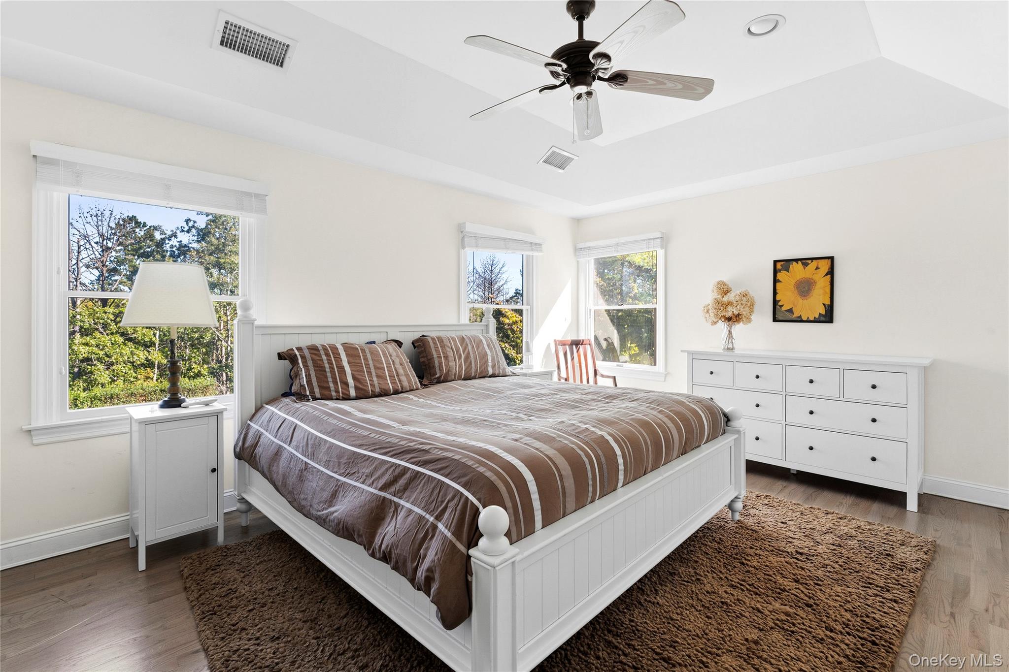 Bedroom with a white bed, brown striped bedding, ceiling fan, and large windows overlooking trees.