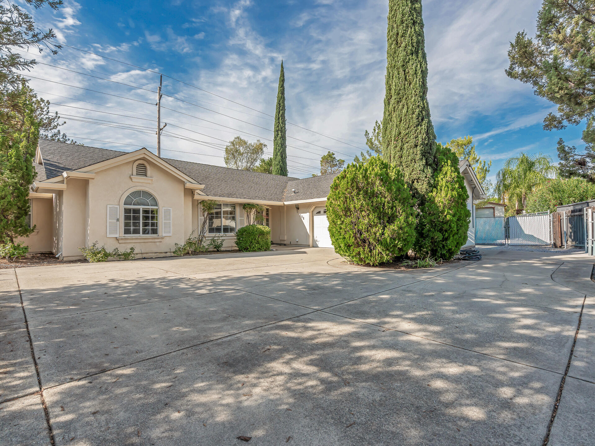 Beige stucco home with an arched window, tall cypress trees, and a wide concrete driveway.