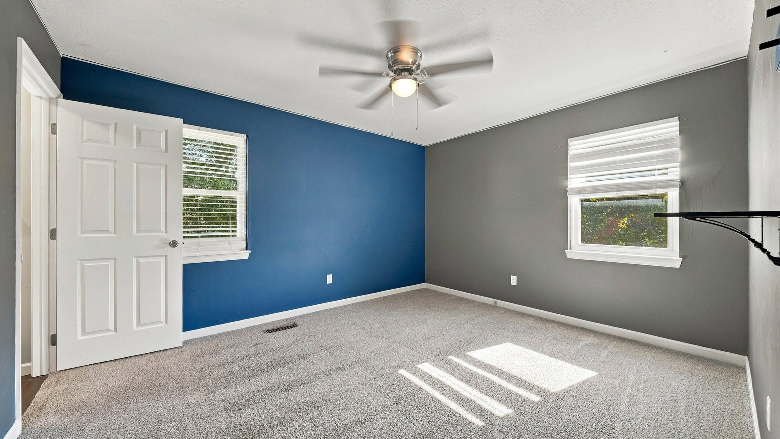 Vacant bedroom with blue accent wall, gray carpet, two windows, and ceiling fan.