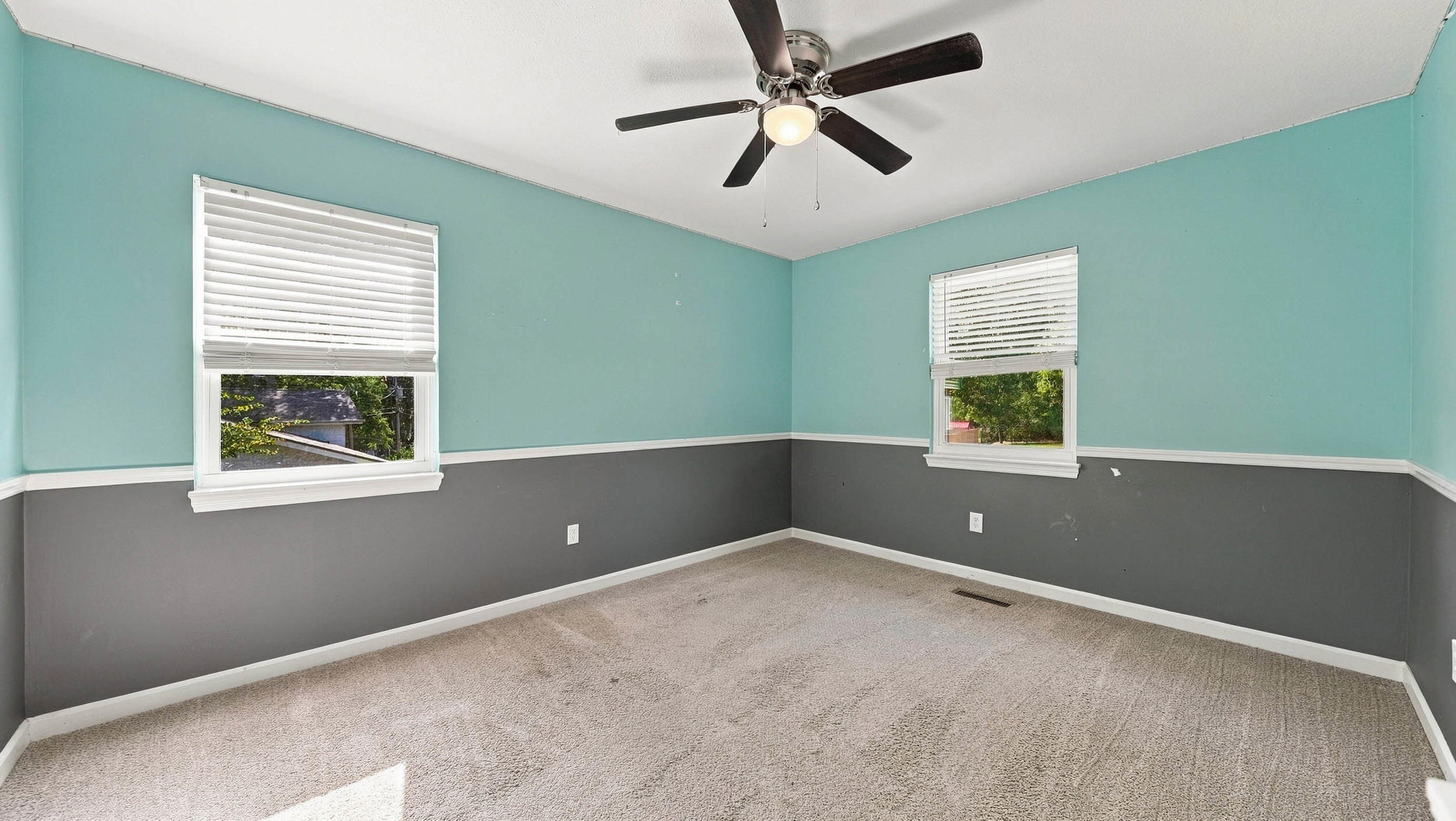 Empty carpeted room with turquoise and gray walls, two windows, and a ceiling fan.