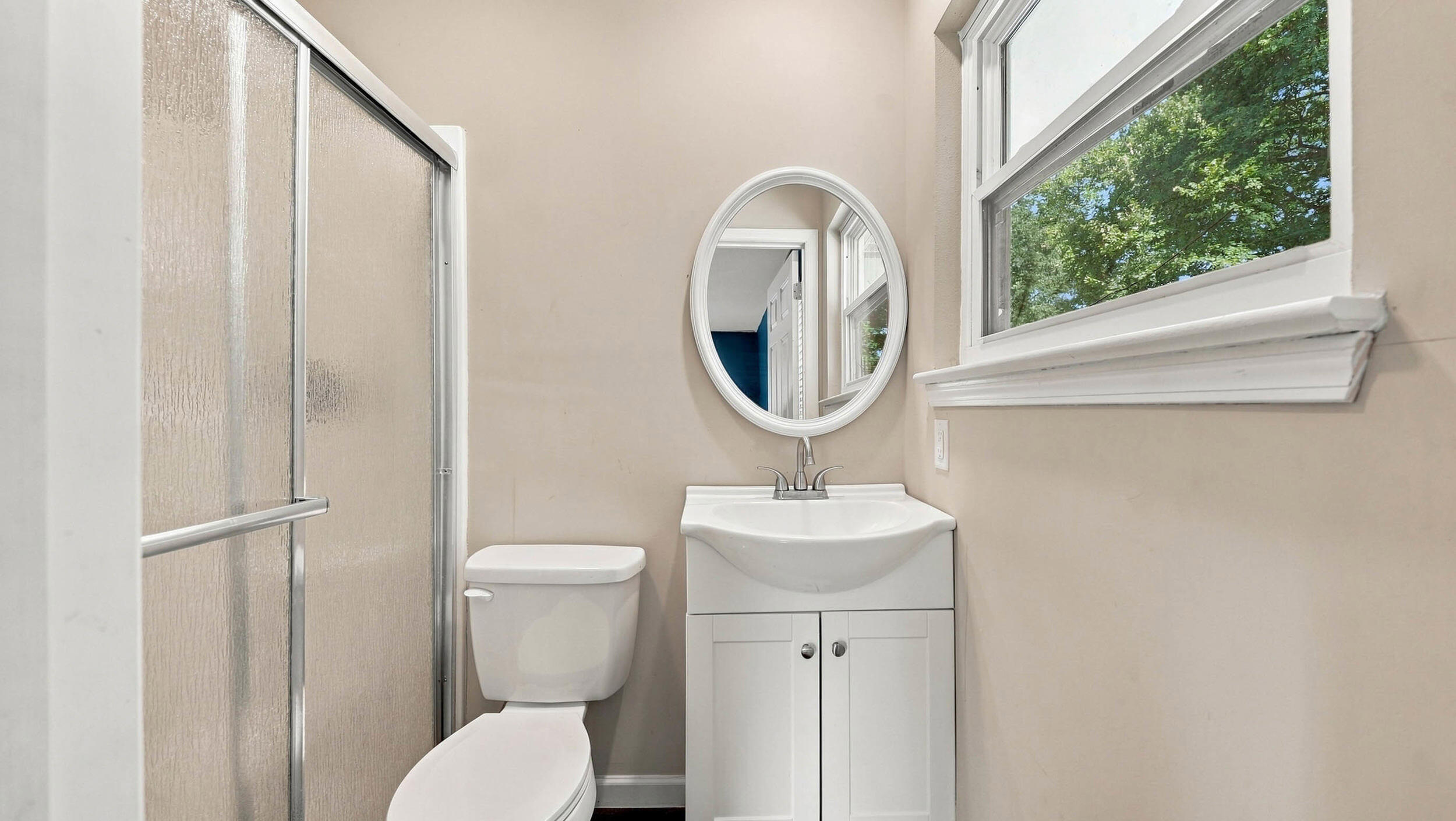 Bathroom with a glass shower, white vanity, oval mirror, and window overlooking trees.