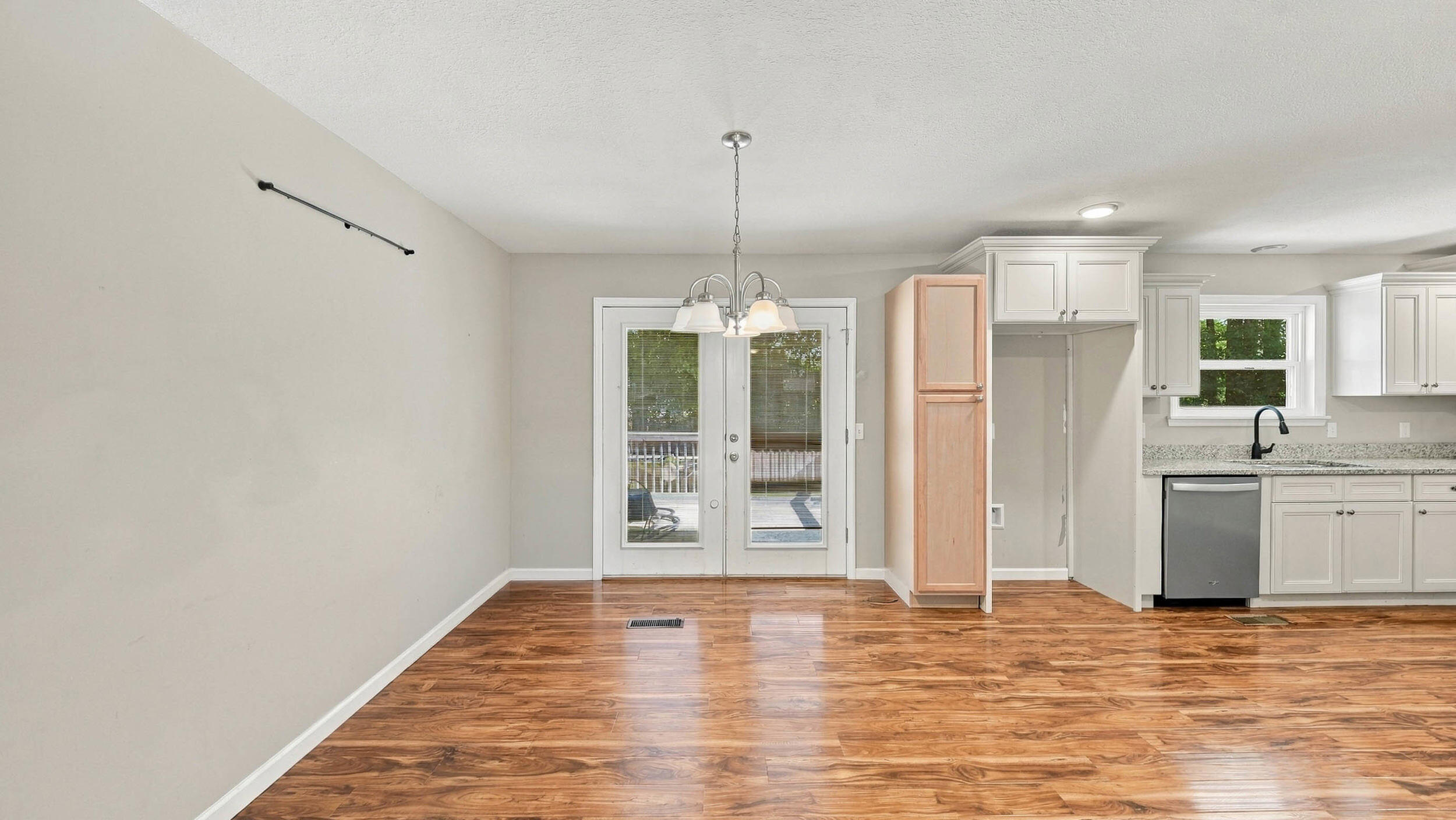 Empty dining area beside a white-cabinet kitchen, with wood-look flooring and glass double doors.