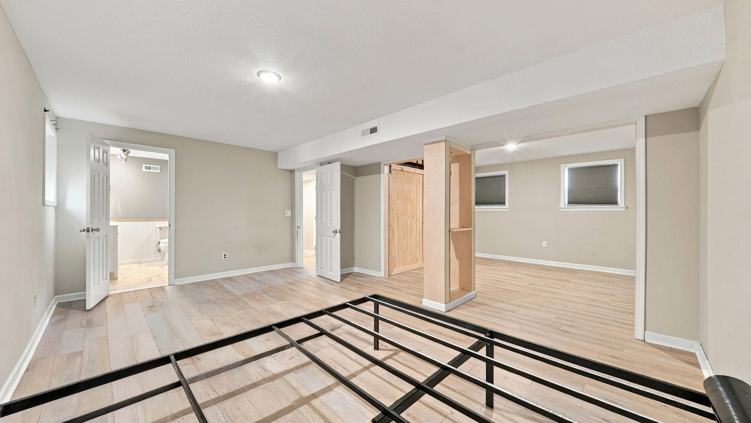 Vacant basement bedroom with an exposed metal bed frame, wood-look floors, and a bathroom doorway.
