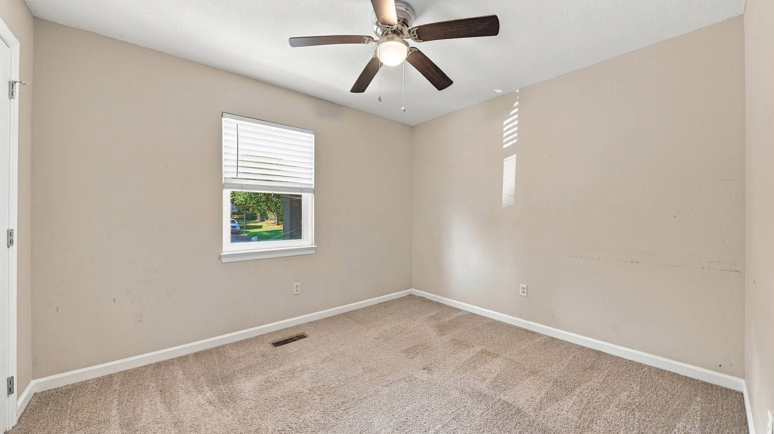Empty carpeted bedroom with beige walls, a window with white blinds, and a ceiling fan.