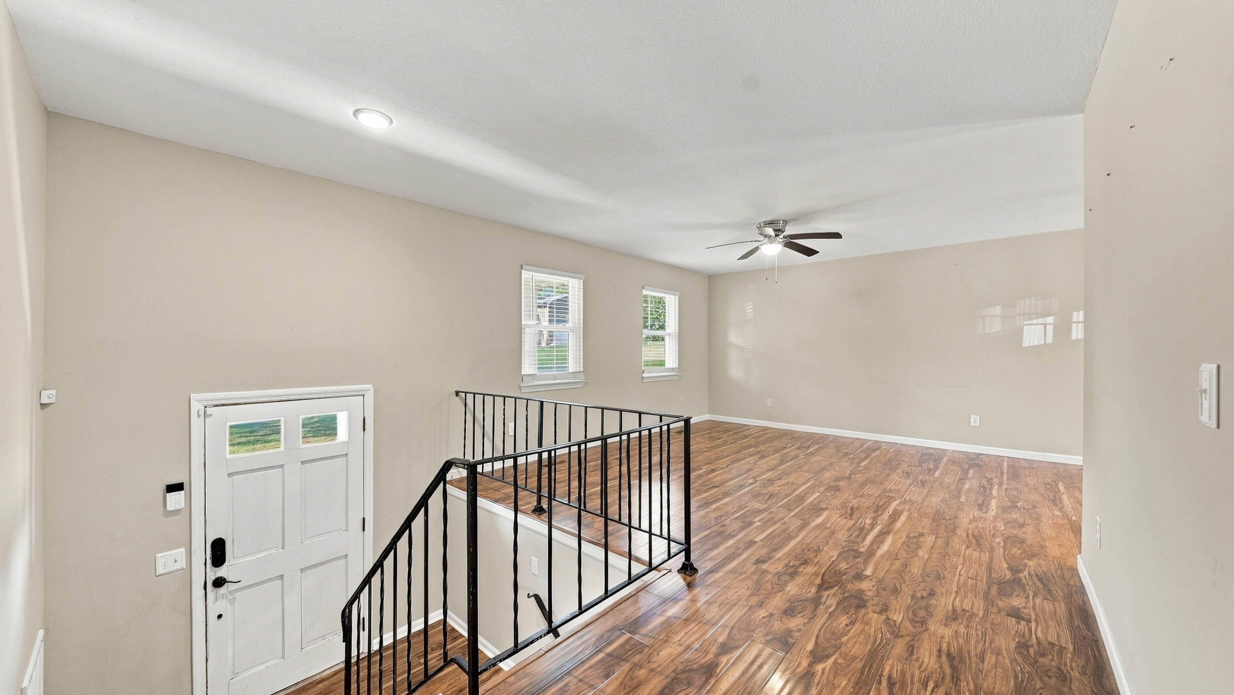 Vacant living area with wood-look floors, ceiling fan, two windows, and a black stair railing.
