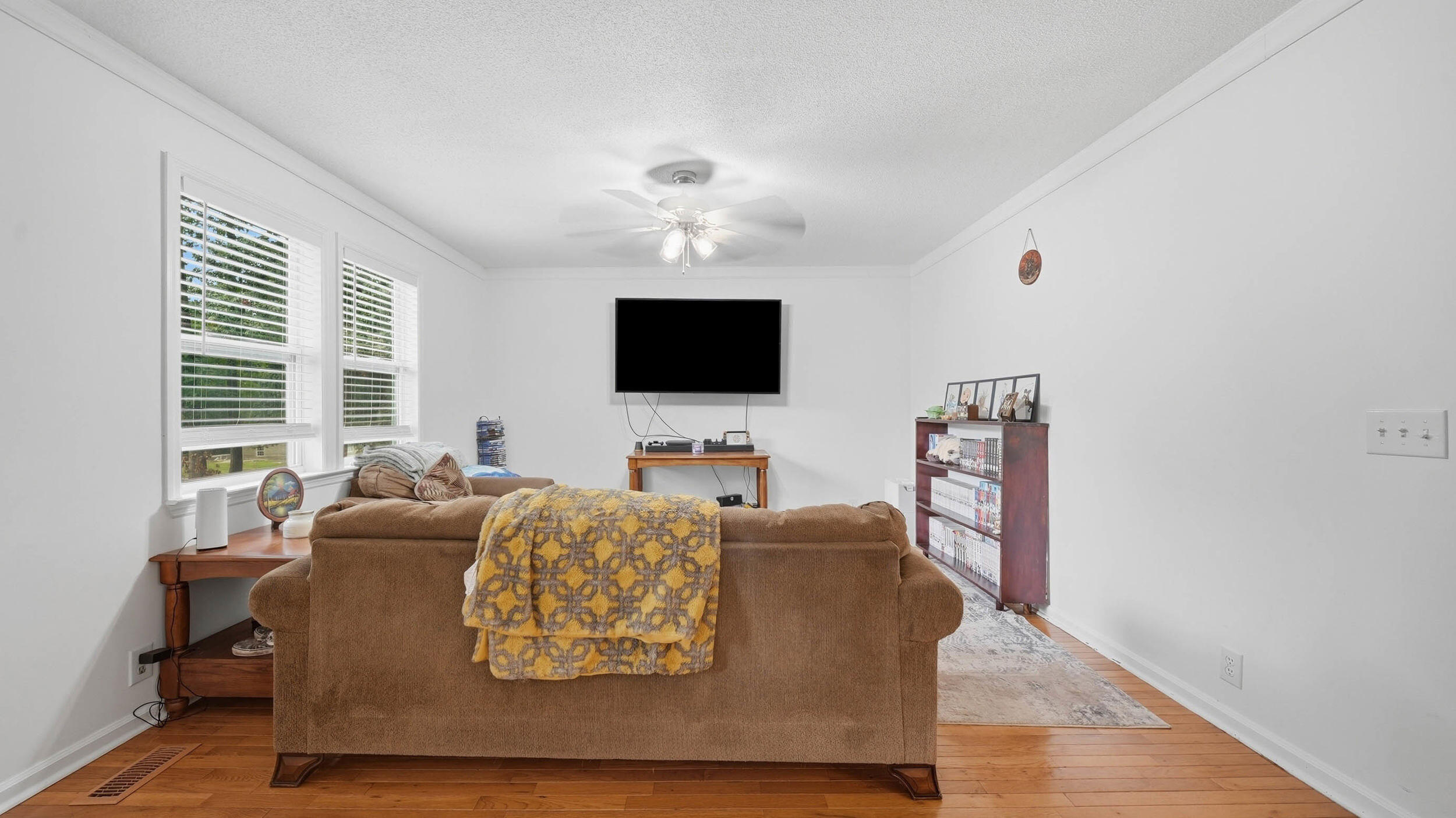 Furnished living room with a brown sofa, yellow patterned throw, wall-mounted TV, and hardwood floors.