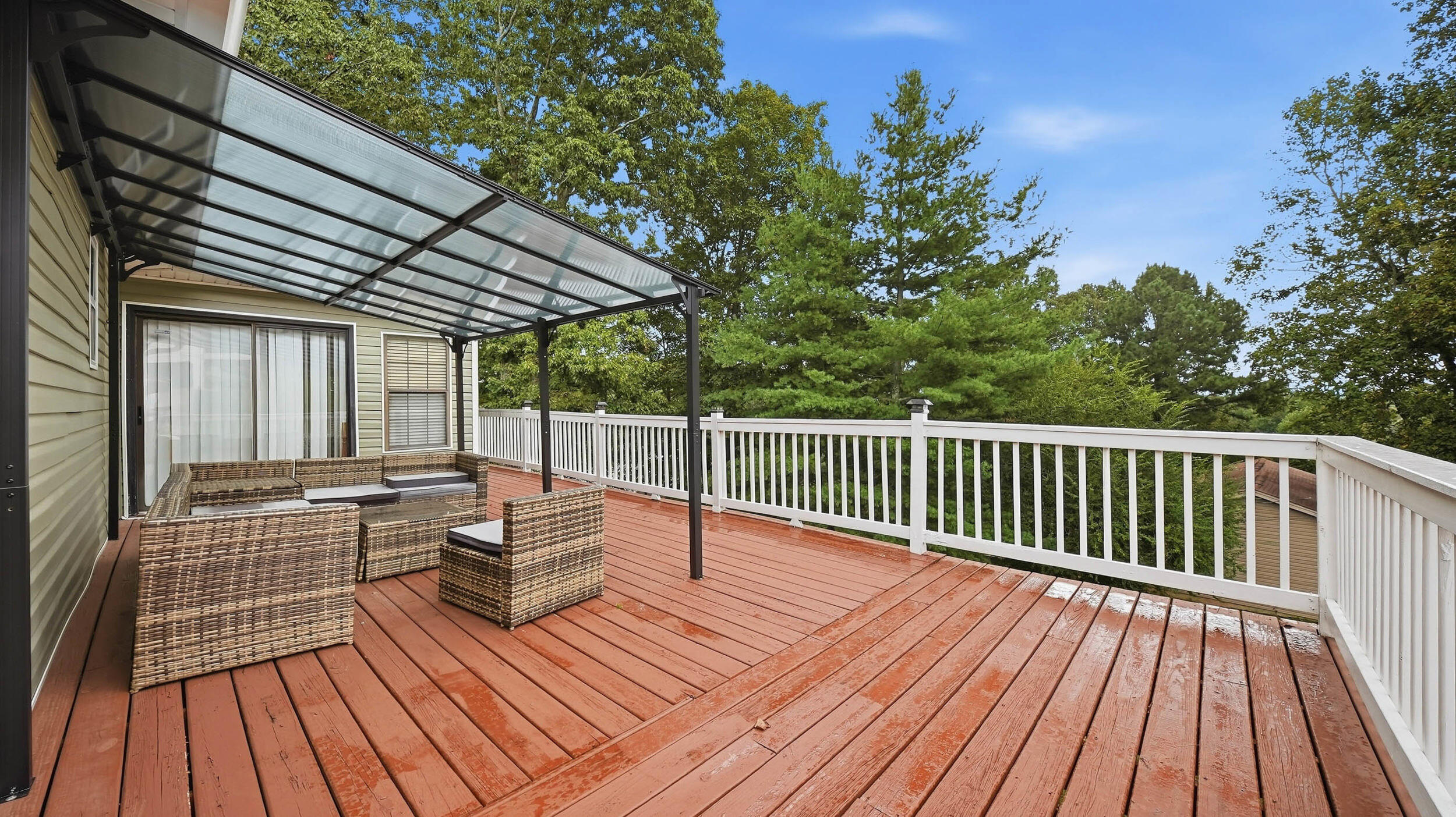 Wood deck with white railings, wicker seating, and a covered patio beside leafy trees.