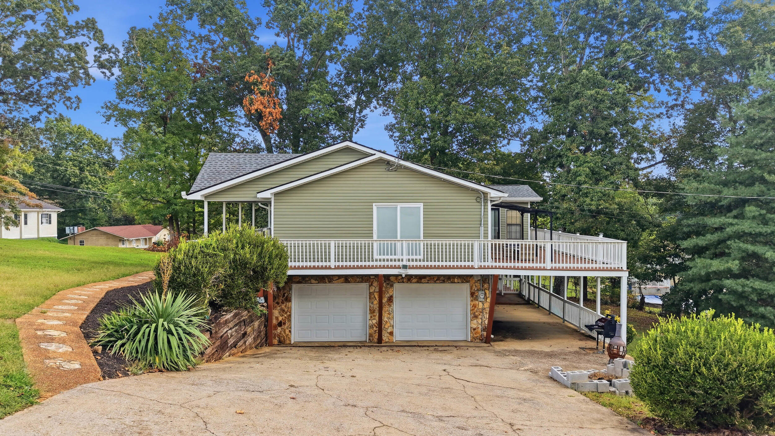 Raised olive-green home with a white-railed deck, two garage doors, and a covered carport.