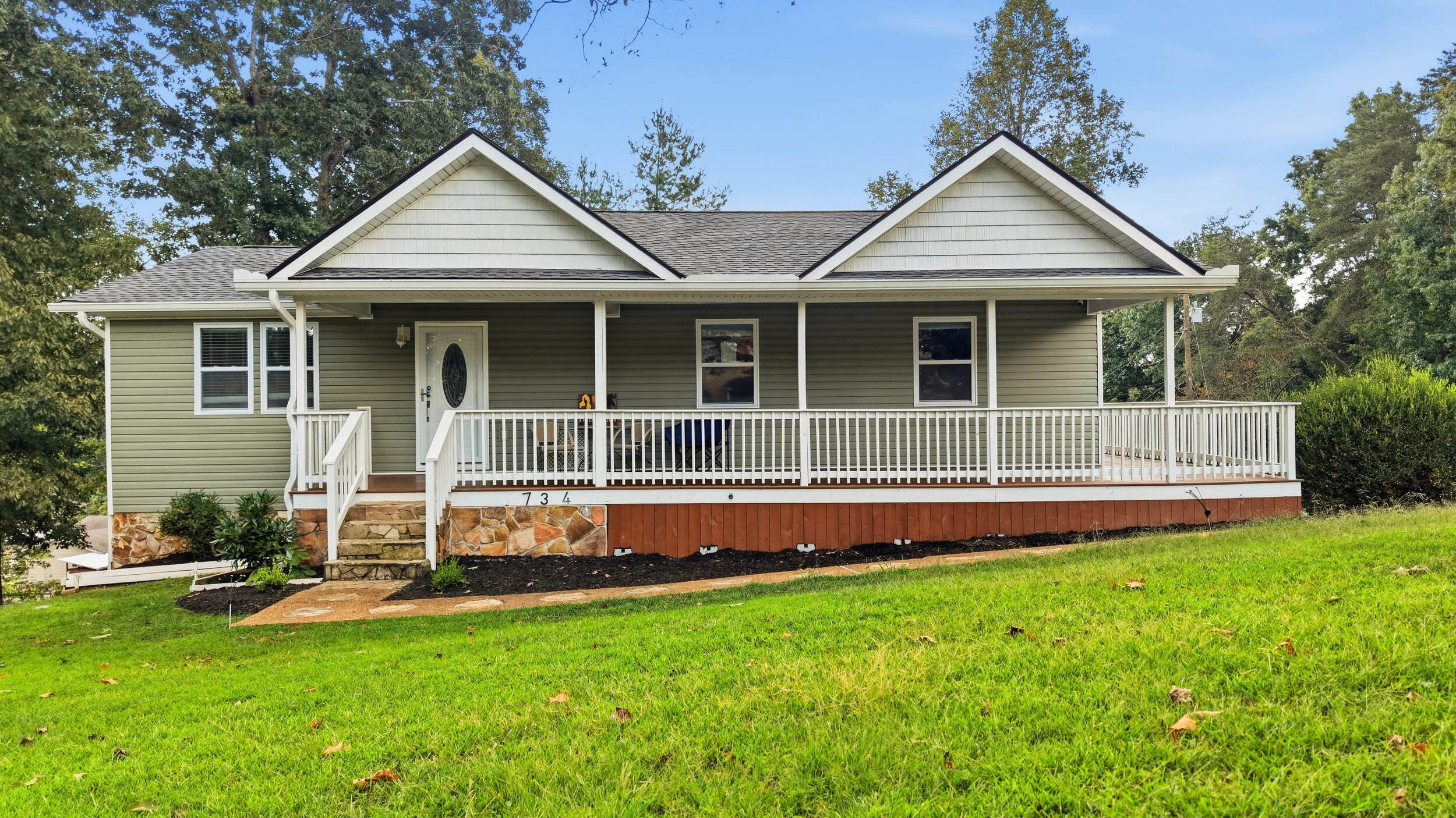 Single-story green-sided home with a long white-railed porch, stone foundation, and broad front lawn.