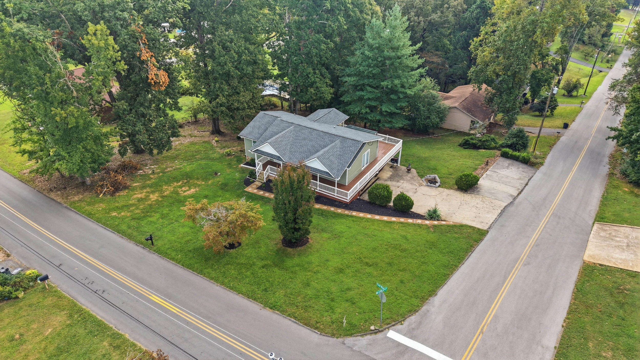 Aerial view of a house with a wraparound porch on a grassy, tree-lined corner lot.