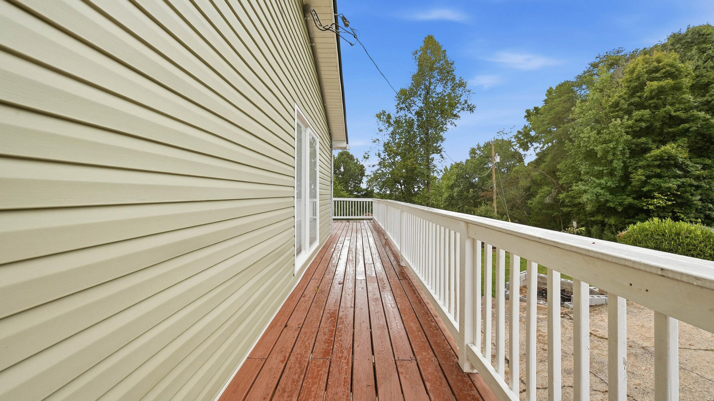 Wood deck with white railings runs beside the home toward a leafy, wooded yard.