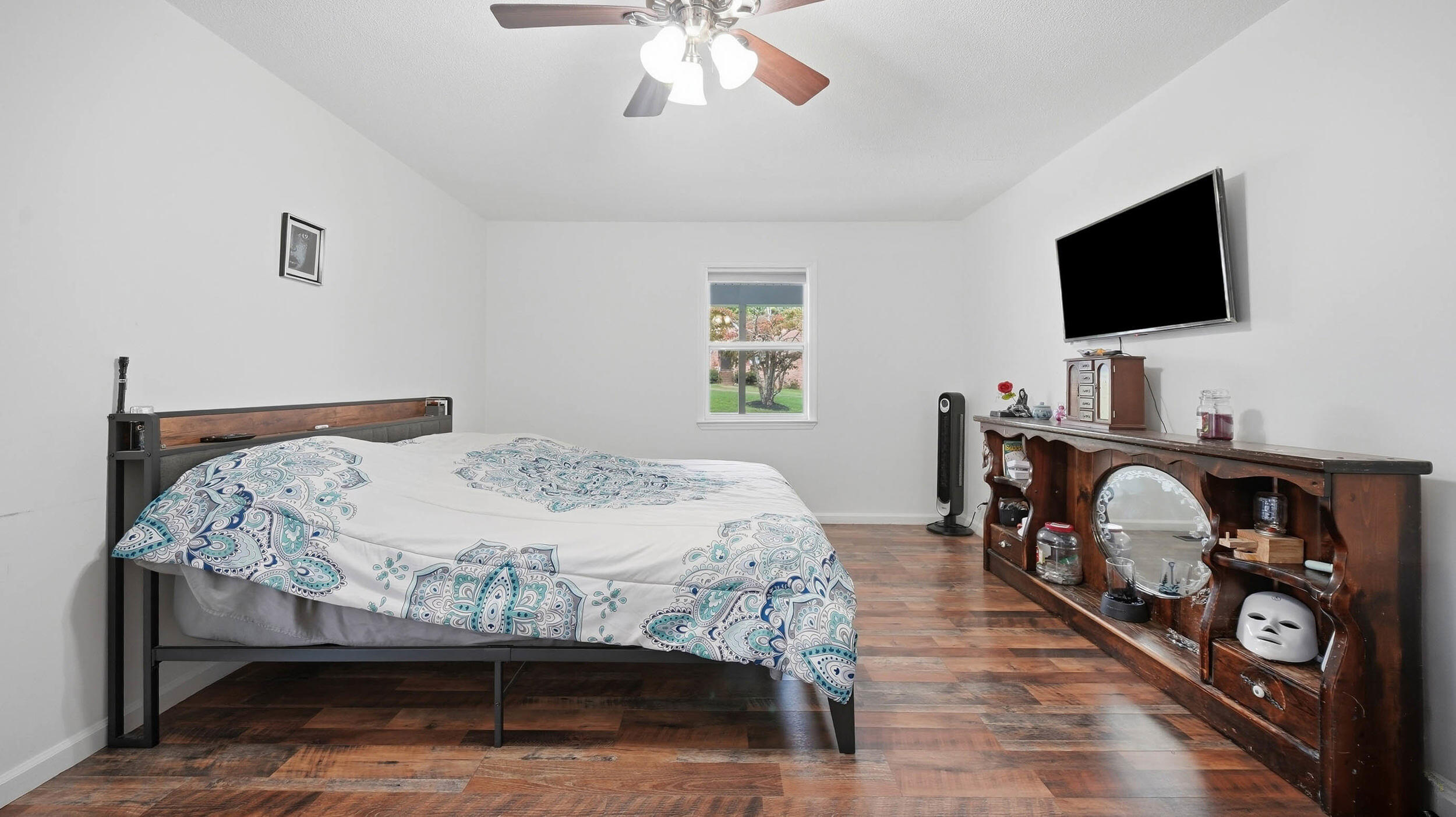 Bedroom with patterned bedding, dark wood floors, and a rustic display cabinet beneath a wall-mounted TV.