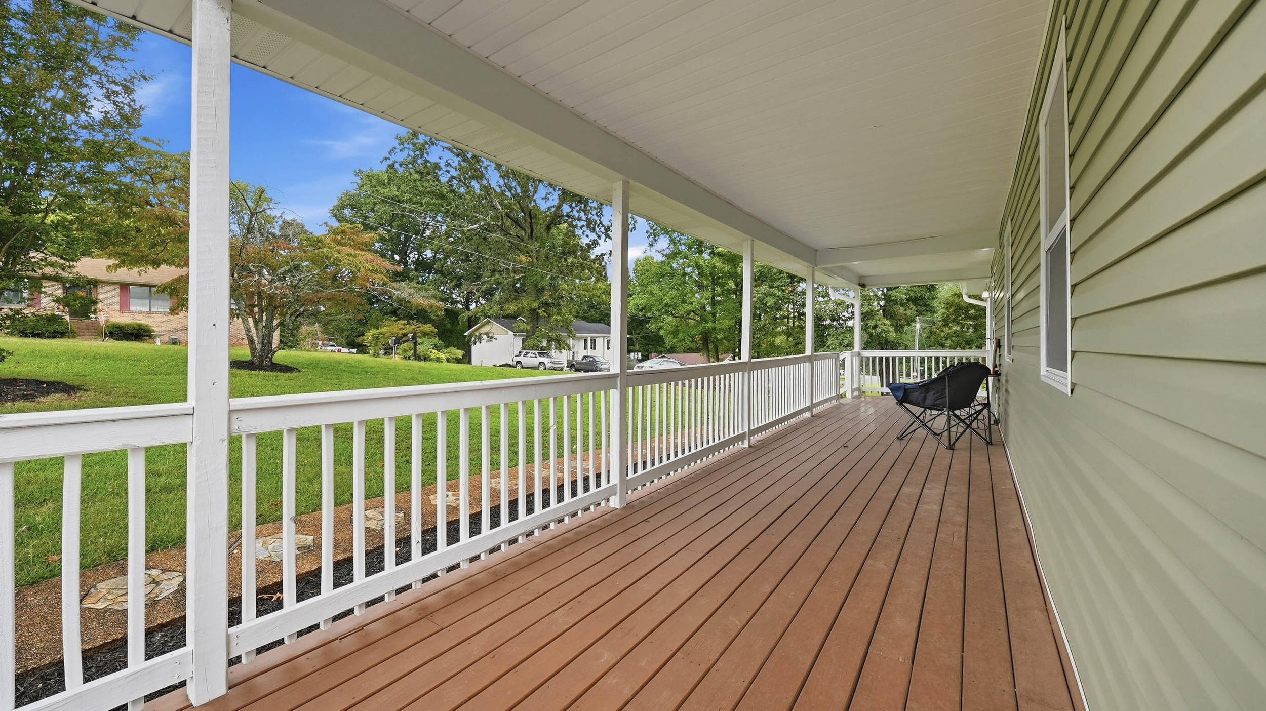 Covered front porch with brown decking, white railings, and black folding chairs overlooking a green lawn.