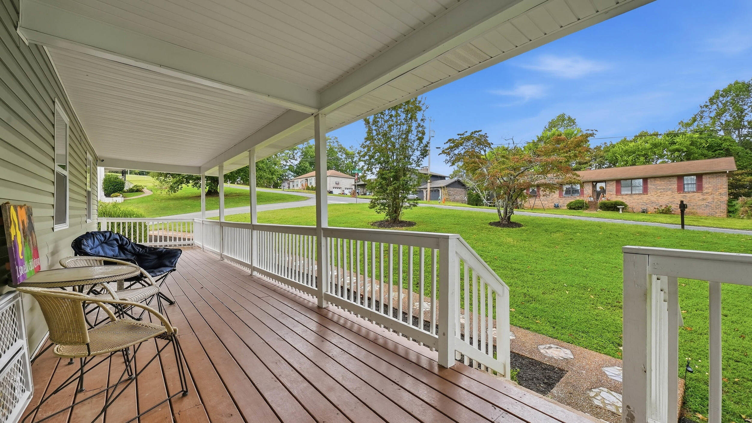 Covered porch with wicker seating and a black lounge chair overlooking a green lawn and neighboring homes.