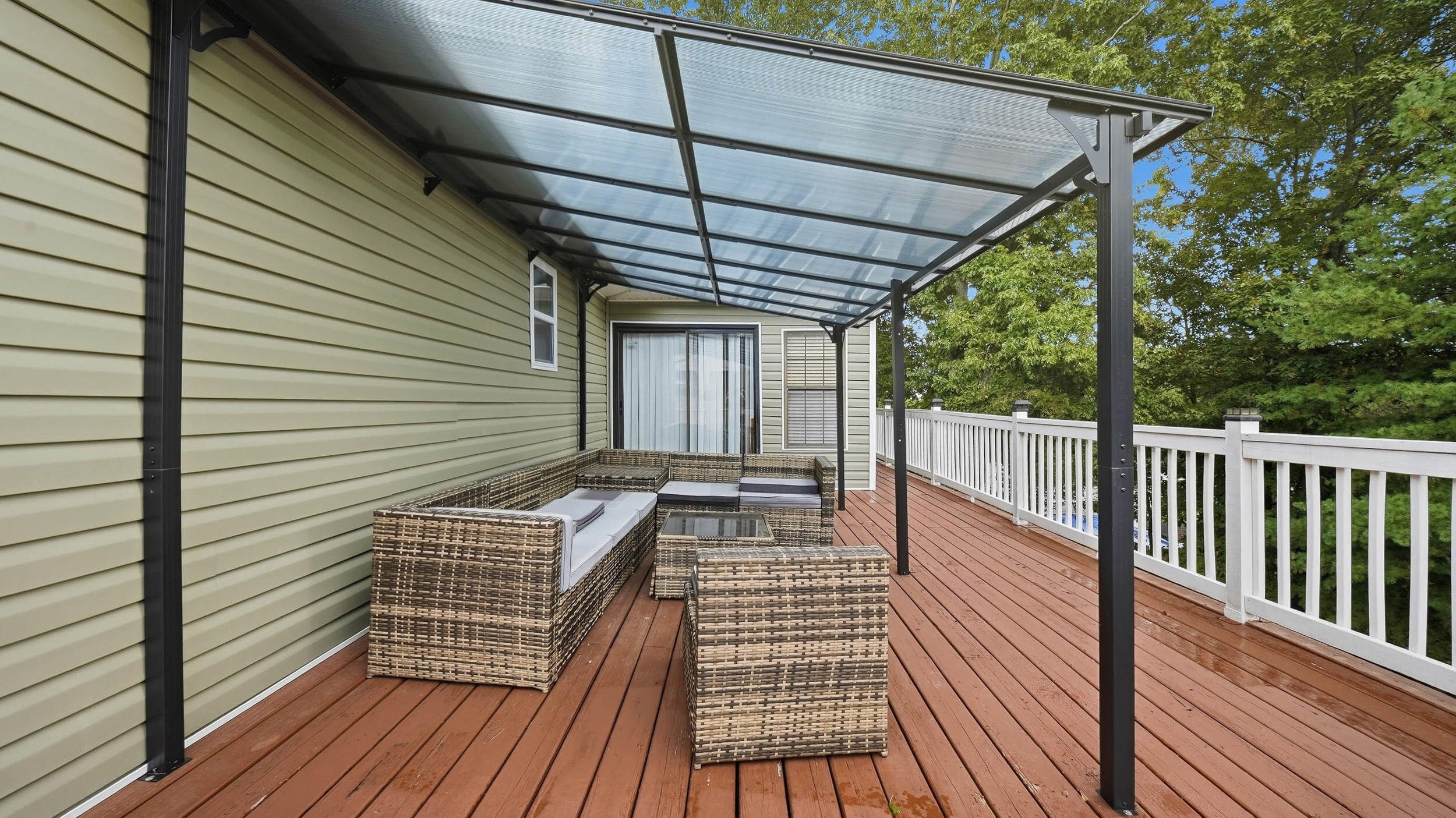 Covered deck with wicker seating, translucent roof, and white railing overlooking trees.