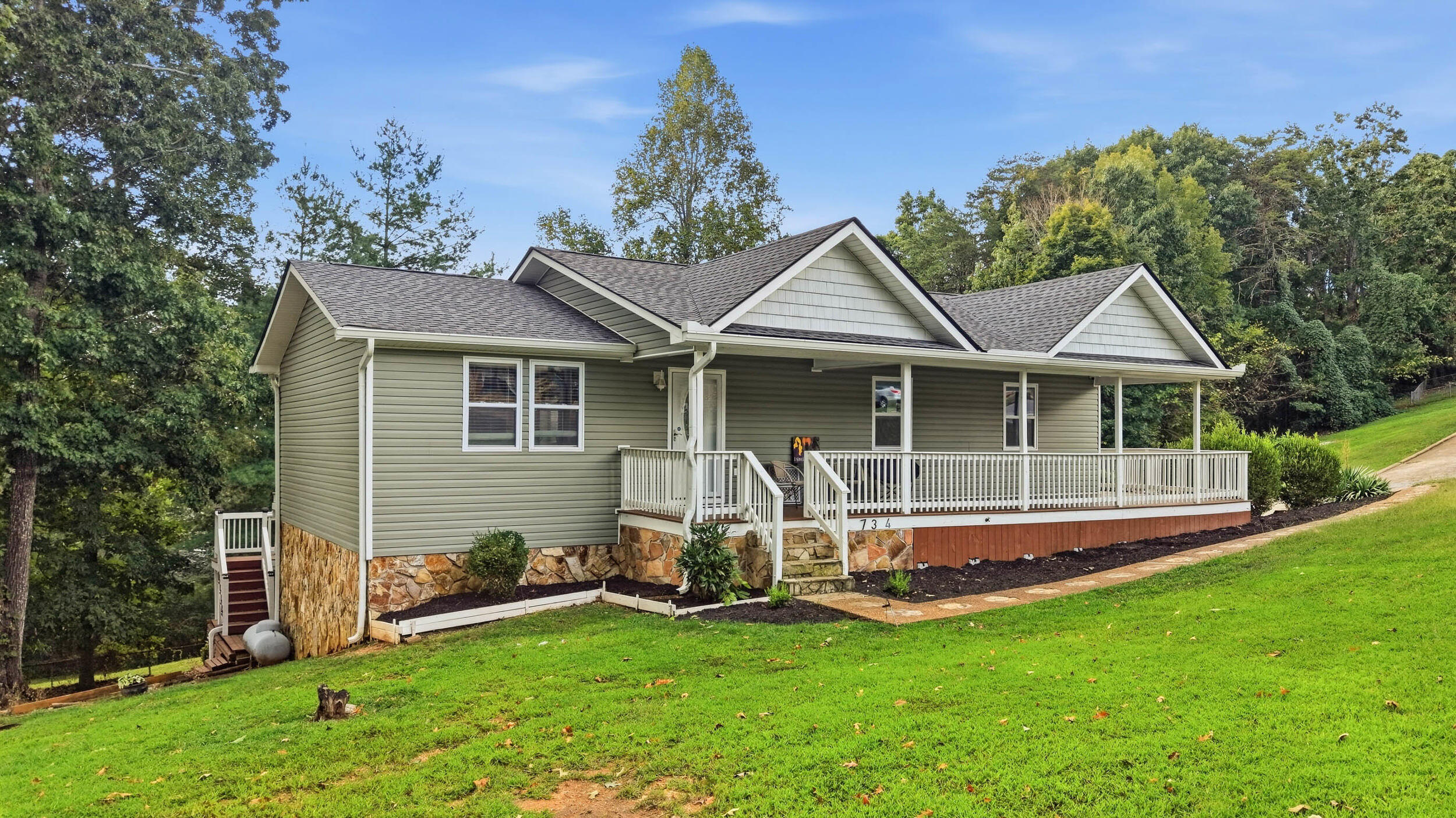 Single-story home with a long covered porch, stone foundation, and sloping green lawn.