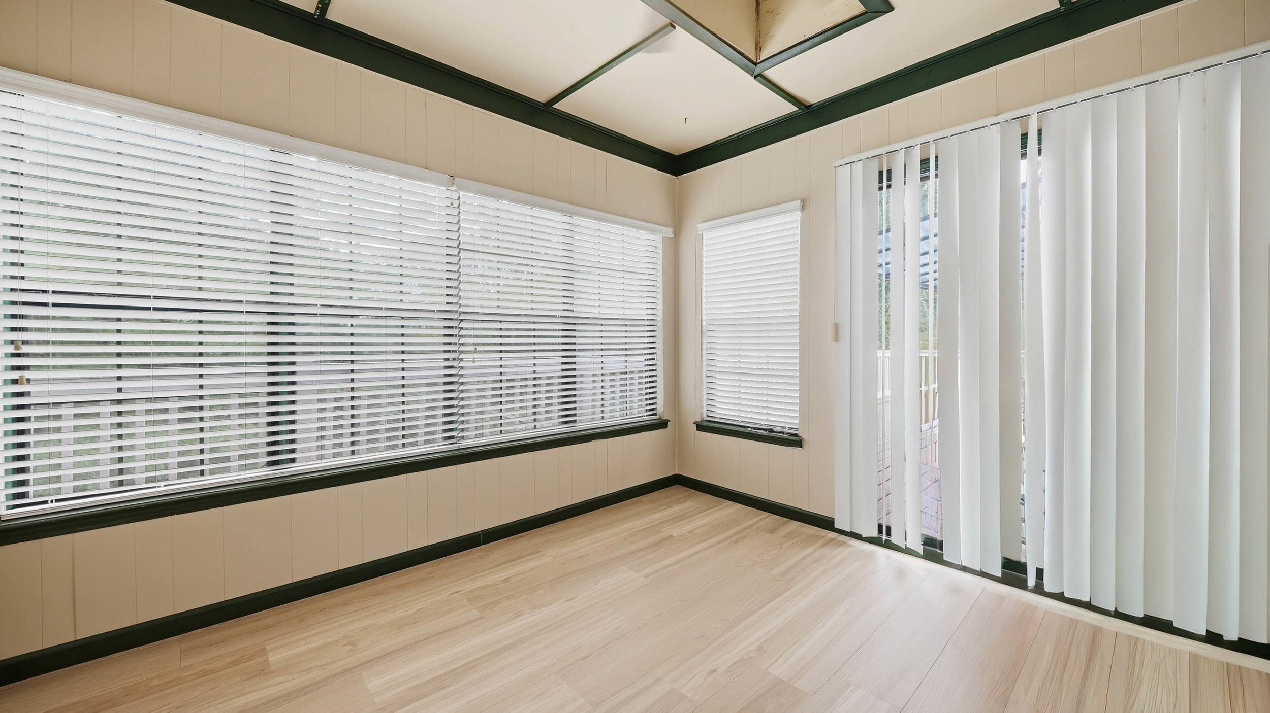 Empty sunroom with light wood flooring, broad windows, and vertical blinds over a sliding glass door.