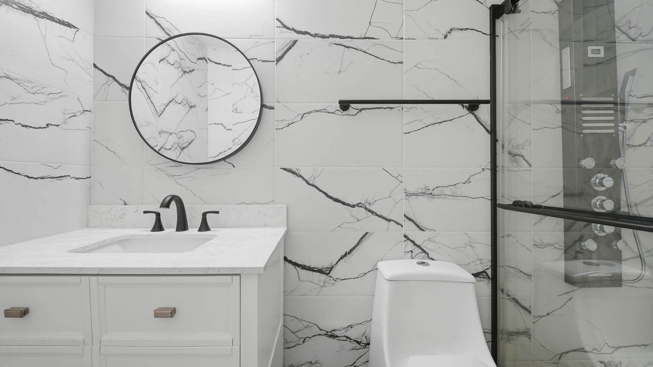 Modern bathroom with veined marble-look tile, a round mirror, white vanity, and glass shower.