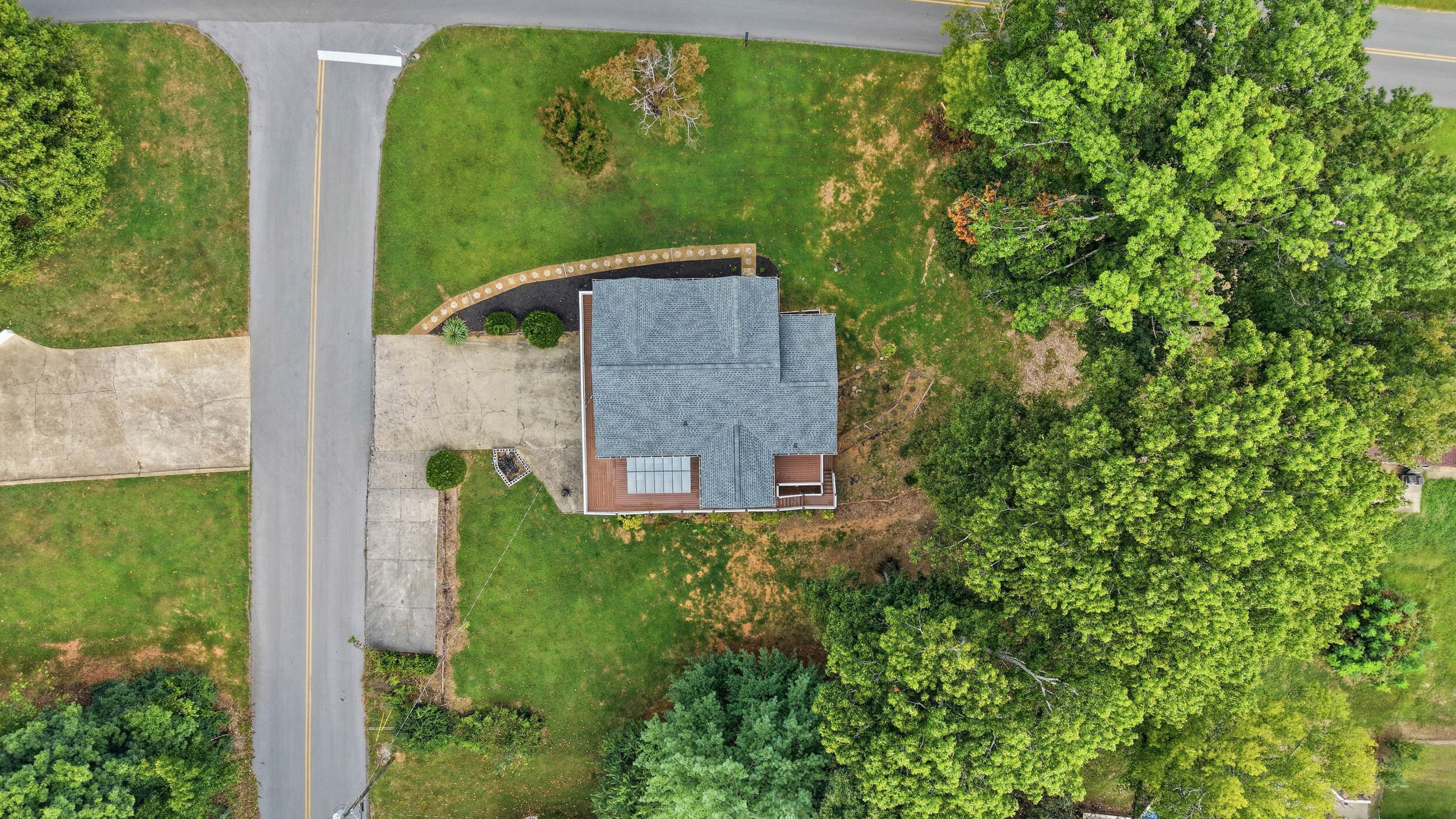 Aerial view of a house, driveway, and lawn beside a road, bordered by mature trees.