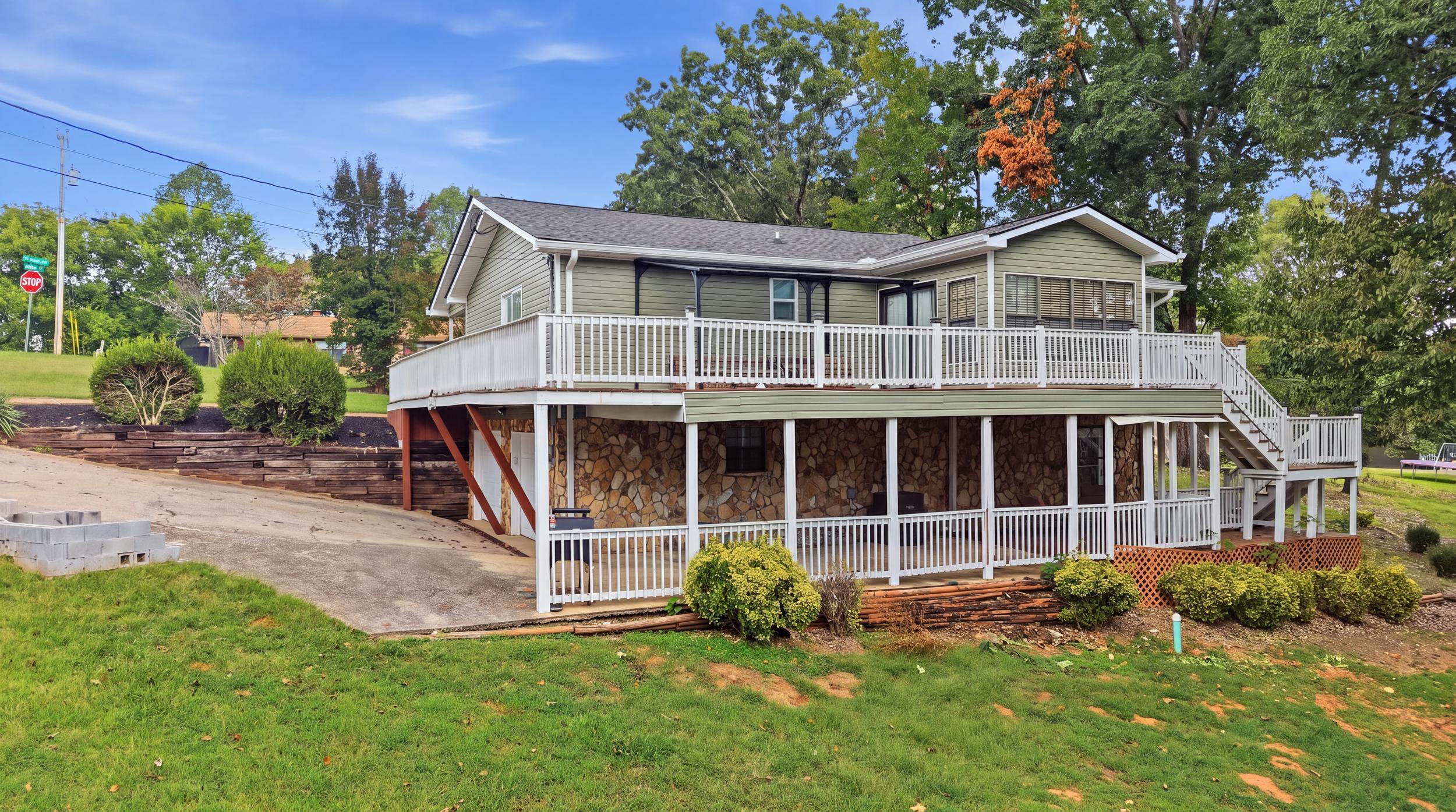 Sage-green home with white-railed decks, a stone lower level, and stairs beside a sloped lawn.