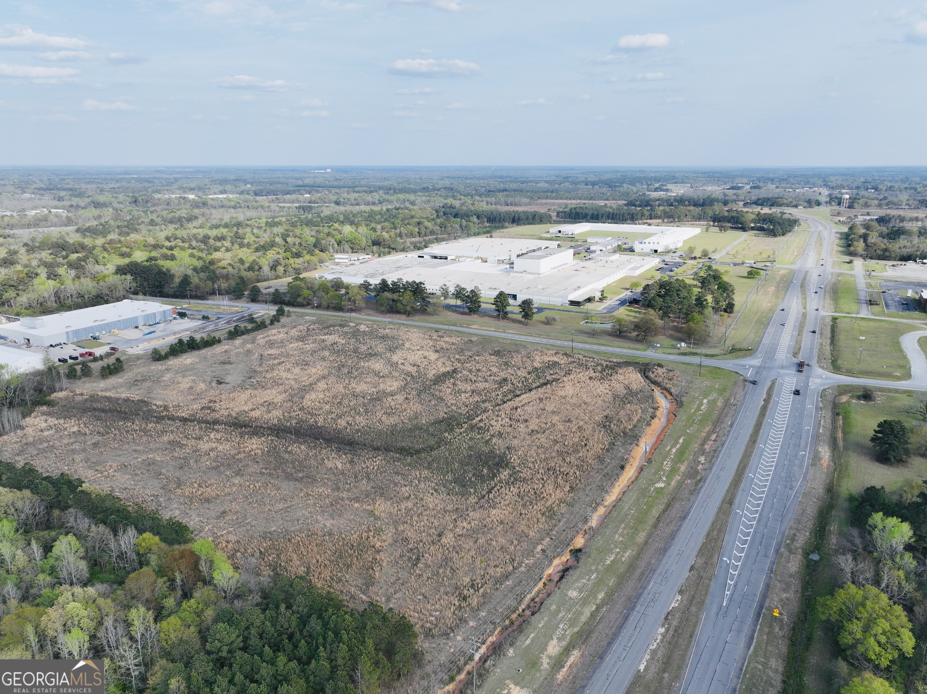 Aerial view of a field beside a divided highway, with industrial buildings and woods beyond.