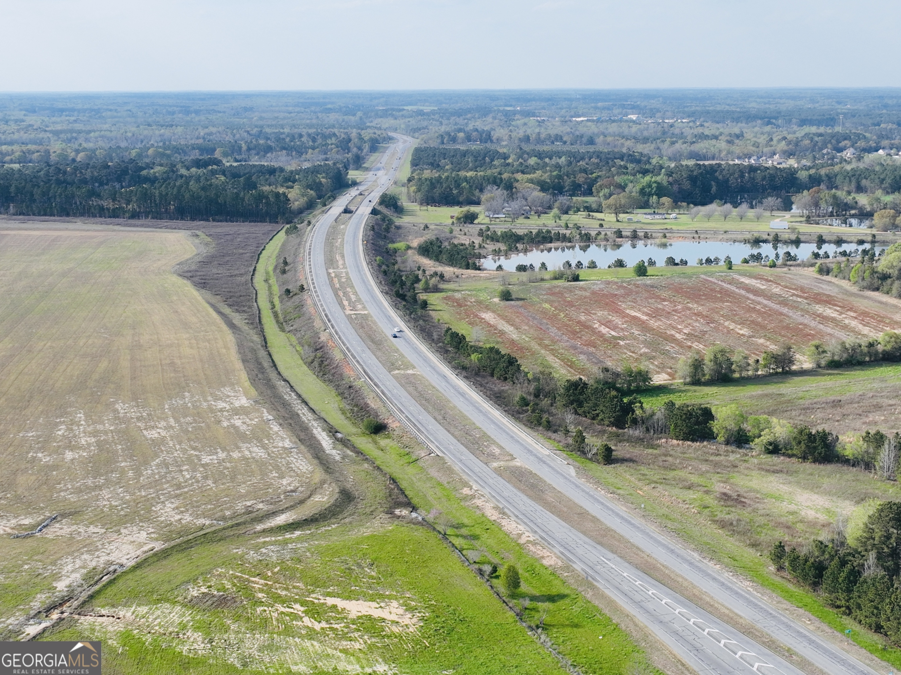 Aerial view of fields beside a divided highway, with a pond and wooded areas nearby.