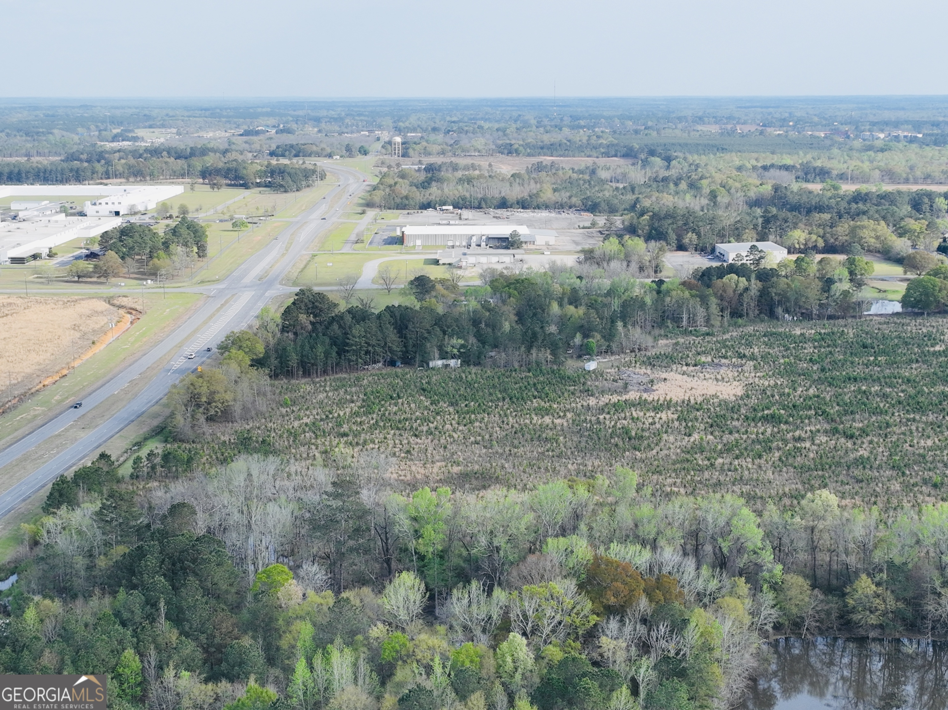 Aerial view of wooded property beside a highway, with industrial buildings and a small pond nearby.