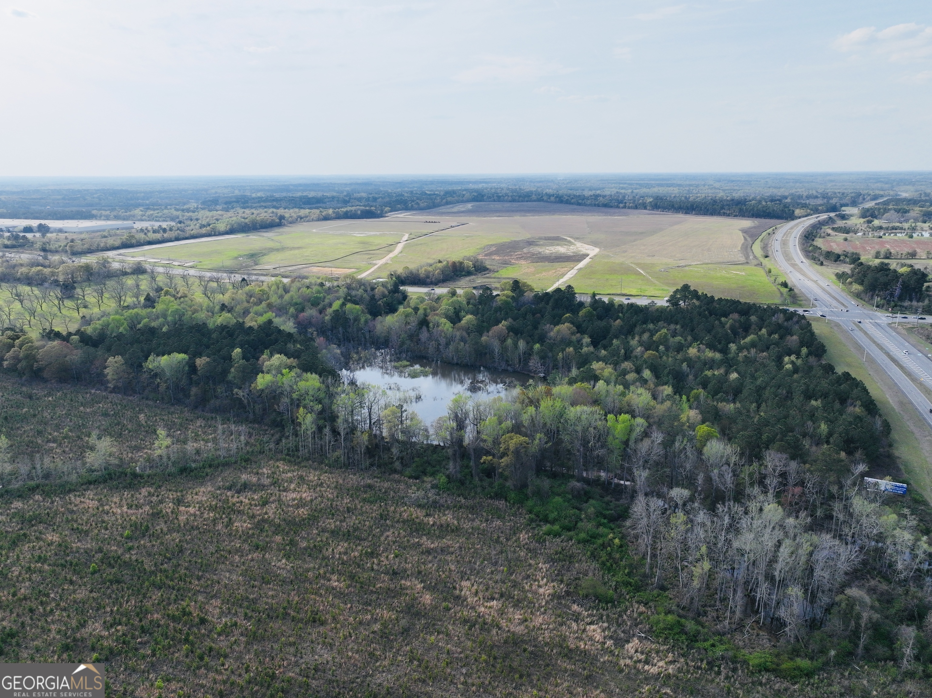 Aerial view of wooded acreage with a pond, open fields, and highway frontage.