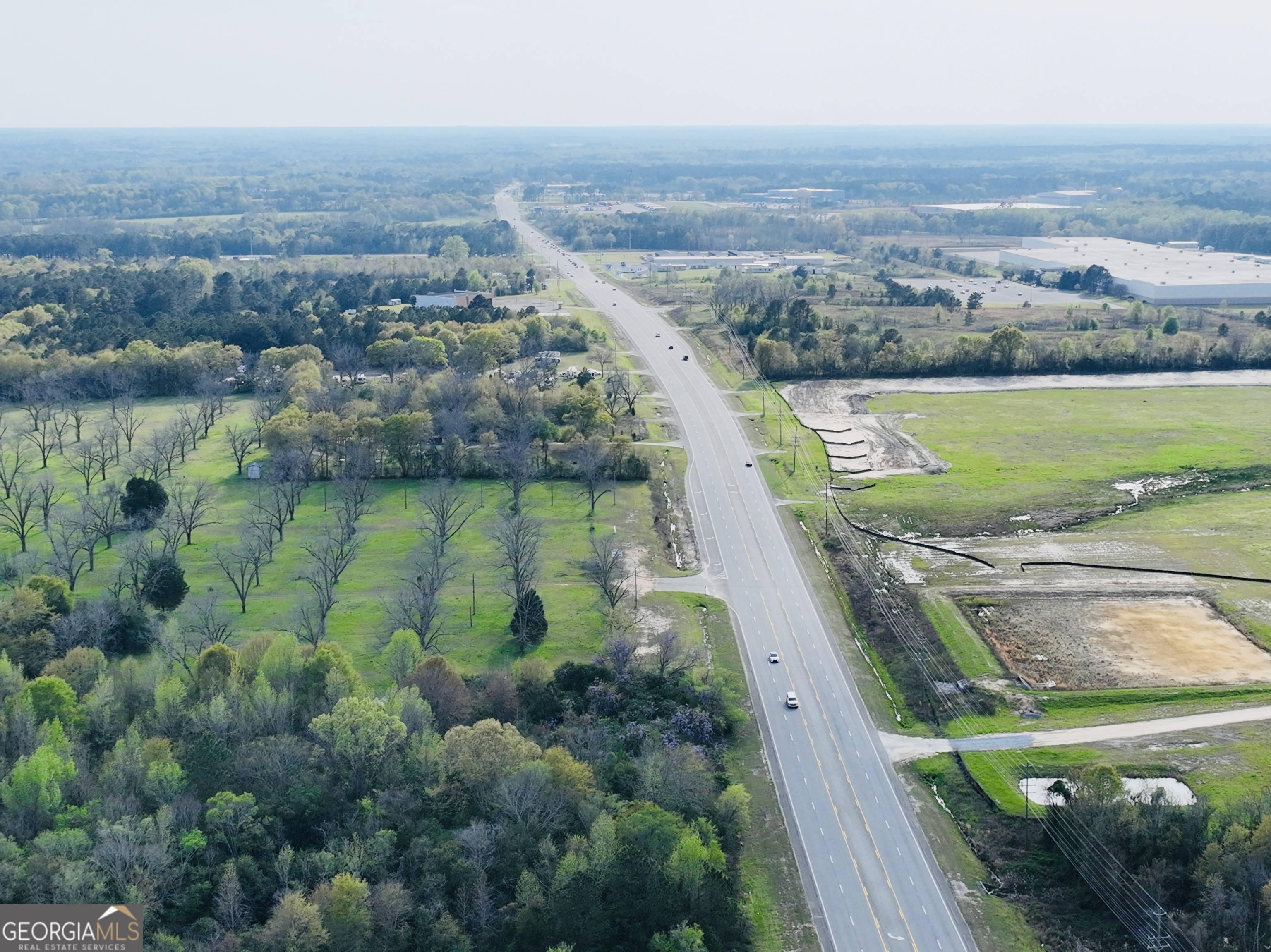 Aerial view of highway frontage beside wooded land, open fields, and nearby commercial buildings.