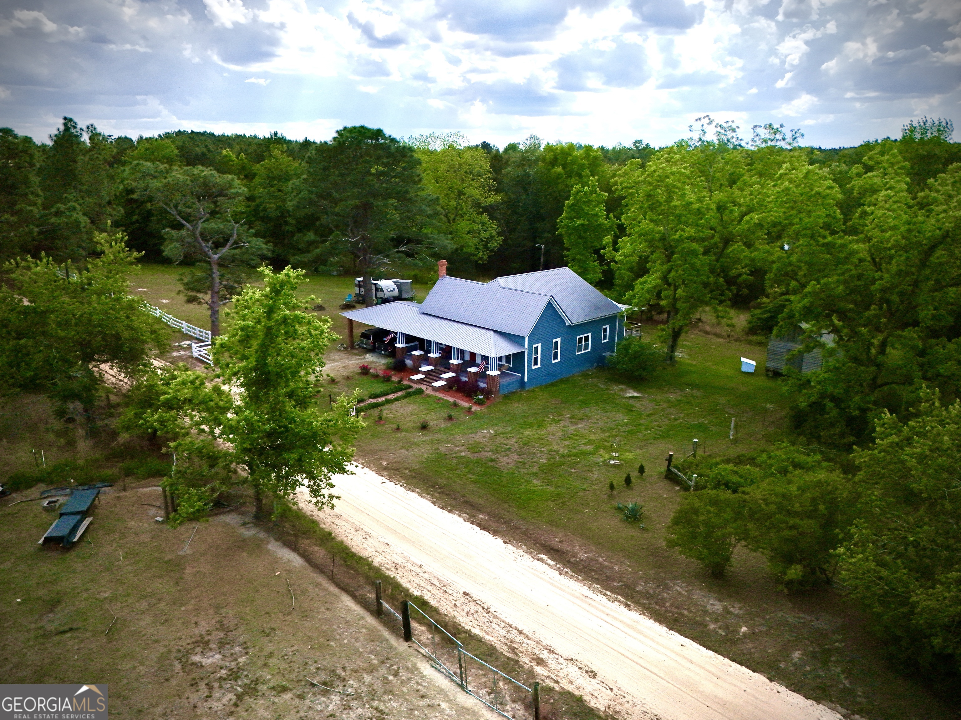 Aerial view of a blue country home with a metal roof, covered porch, gravel drive, and surrounding trees.