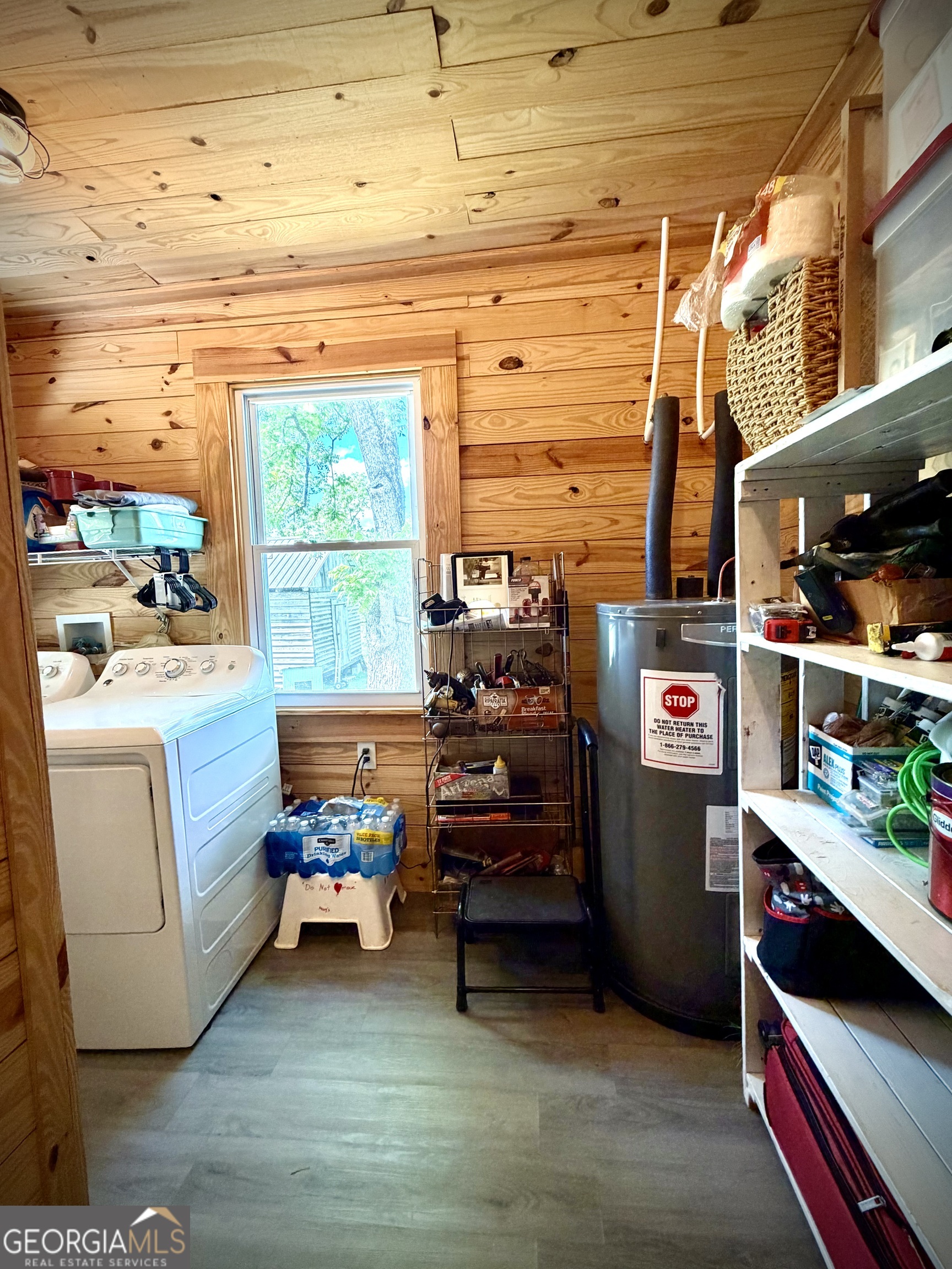 Wood-paneled laundry room with a washer, dryer, water heater, and crowded storage shelves.