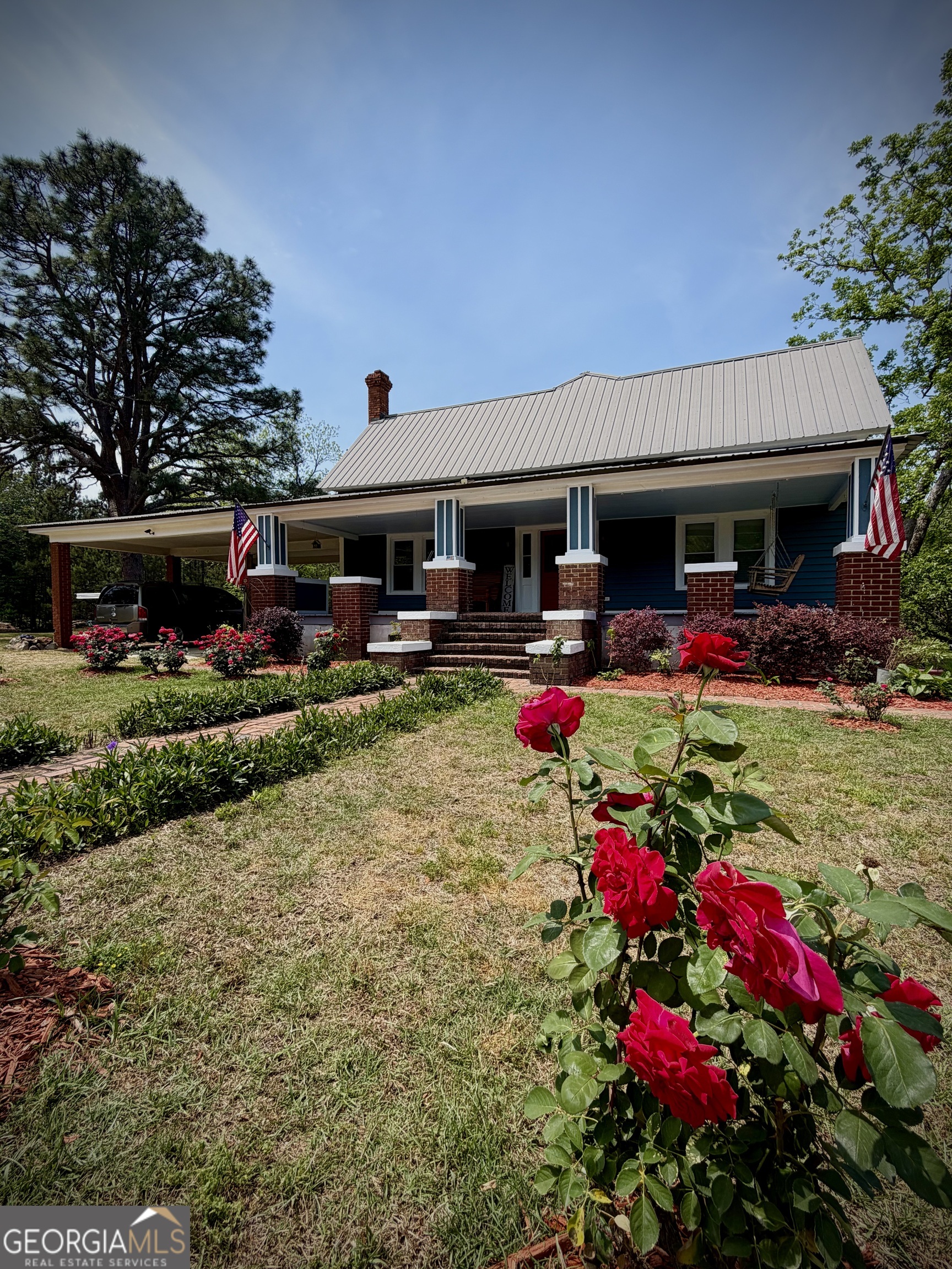Blue house with a brick-columned porch, American flags, and red roses in the foreground.