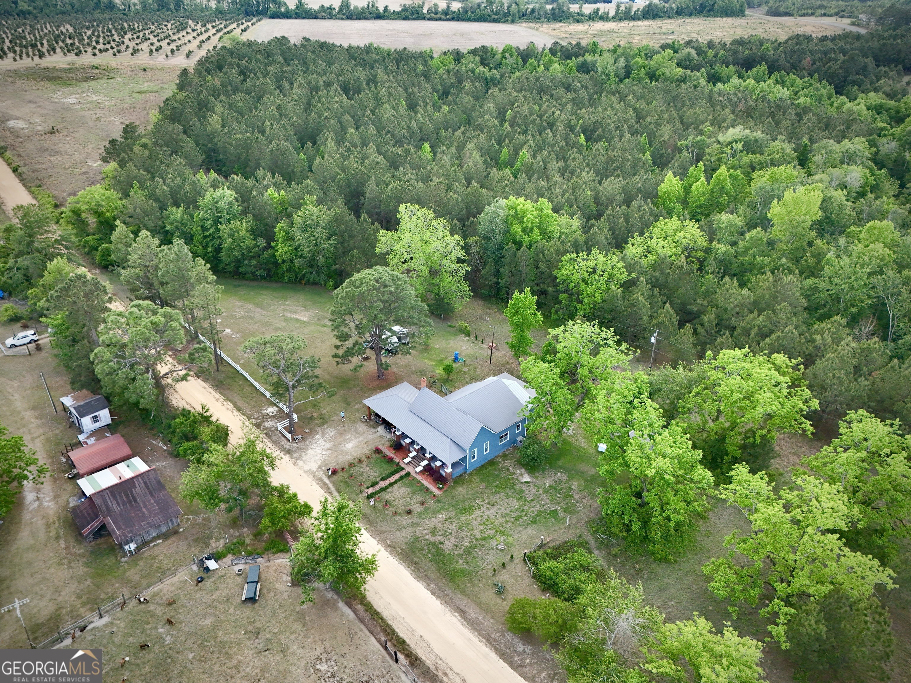 Aerial view of a blue rural home and outbuildings beside a dirt road, surrounded by woods.