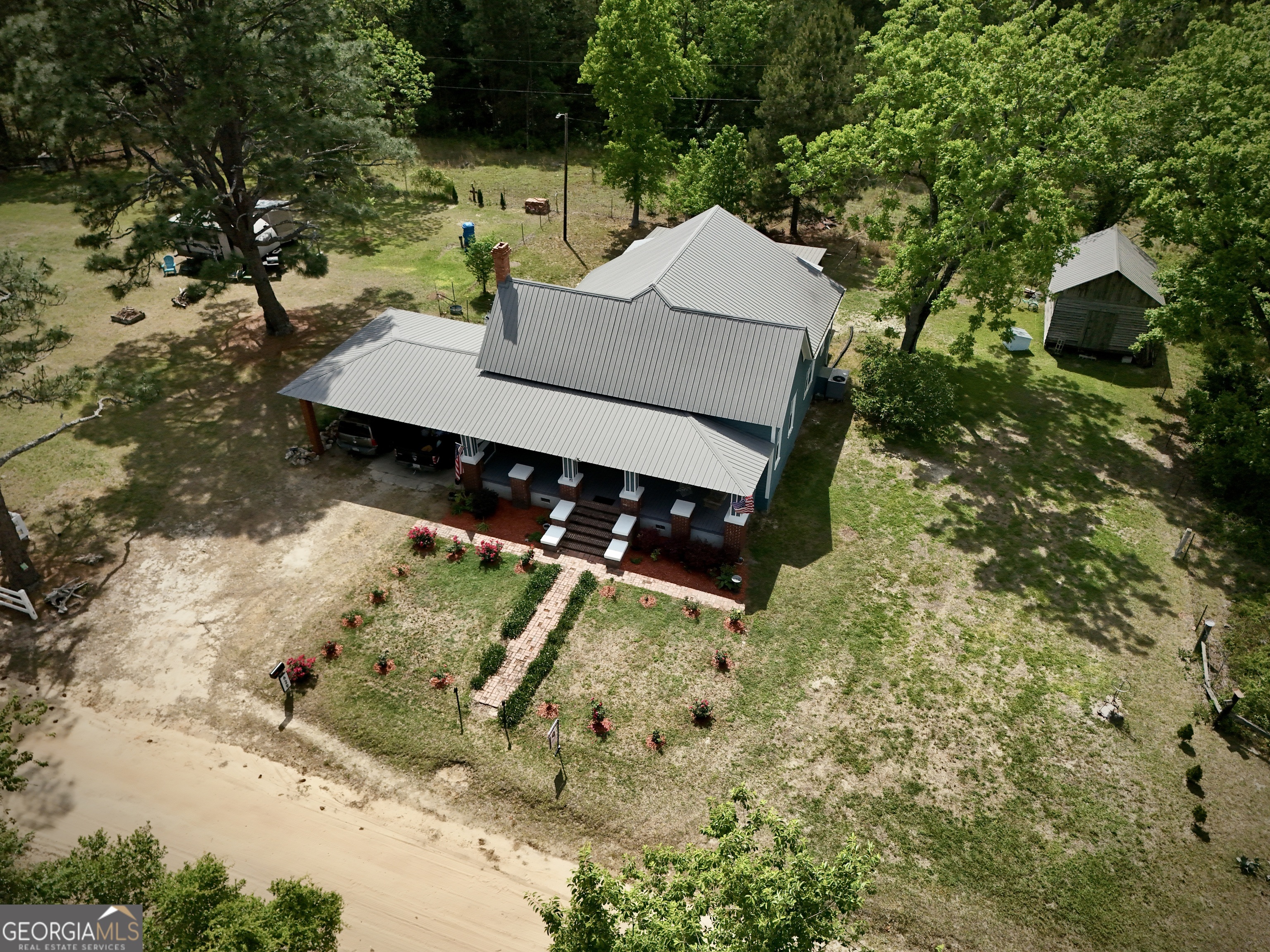 Aerial view of a gray metal-roofed home with a covered carport, brick walkway, and detached shed.
