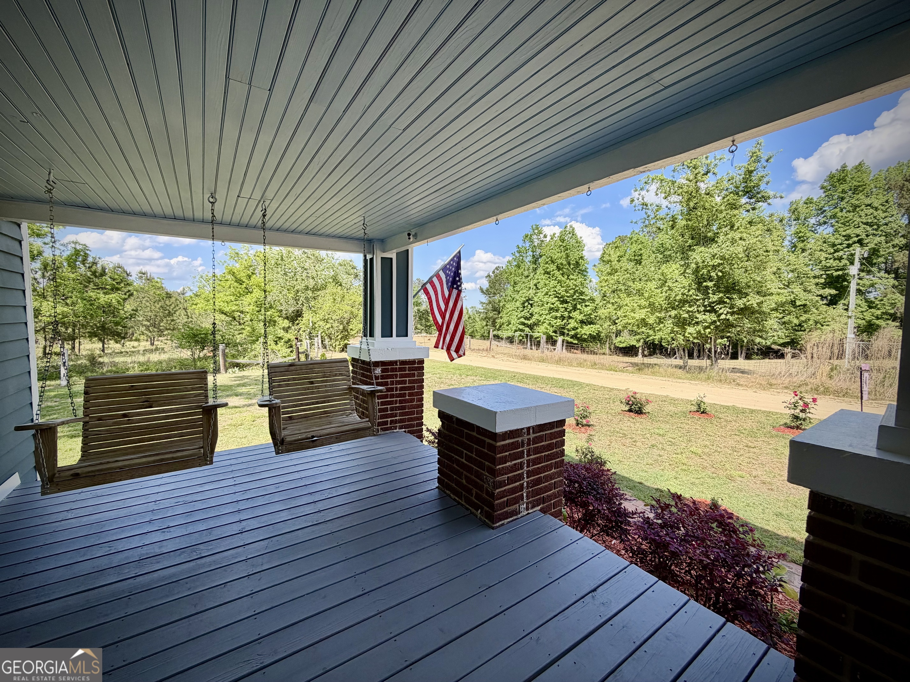 Covered porch with two wooden swings and an American flag overlooking a sunny lawn and trees.