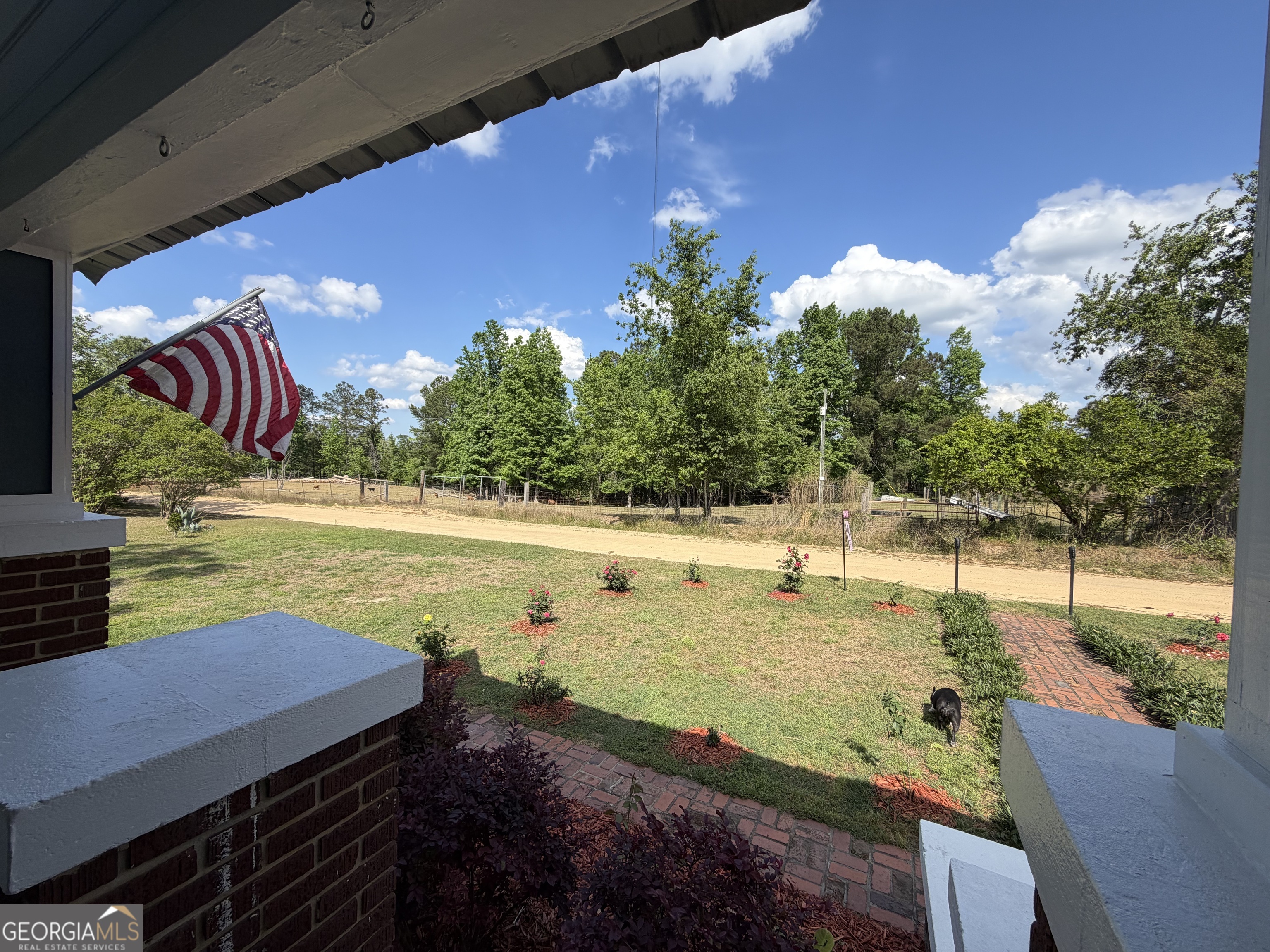 Covered porch overlooks a landscaped yard and rural dirt road, with an American flag and a black cat.
