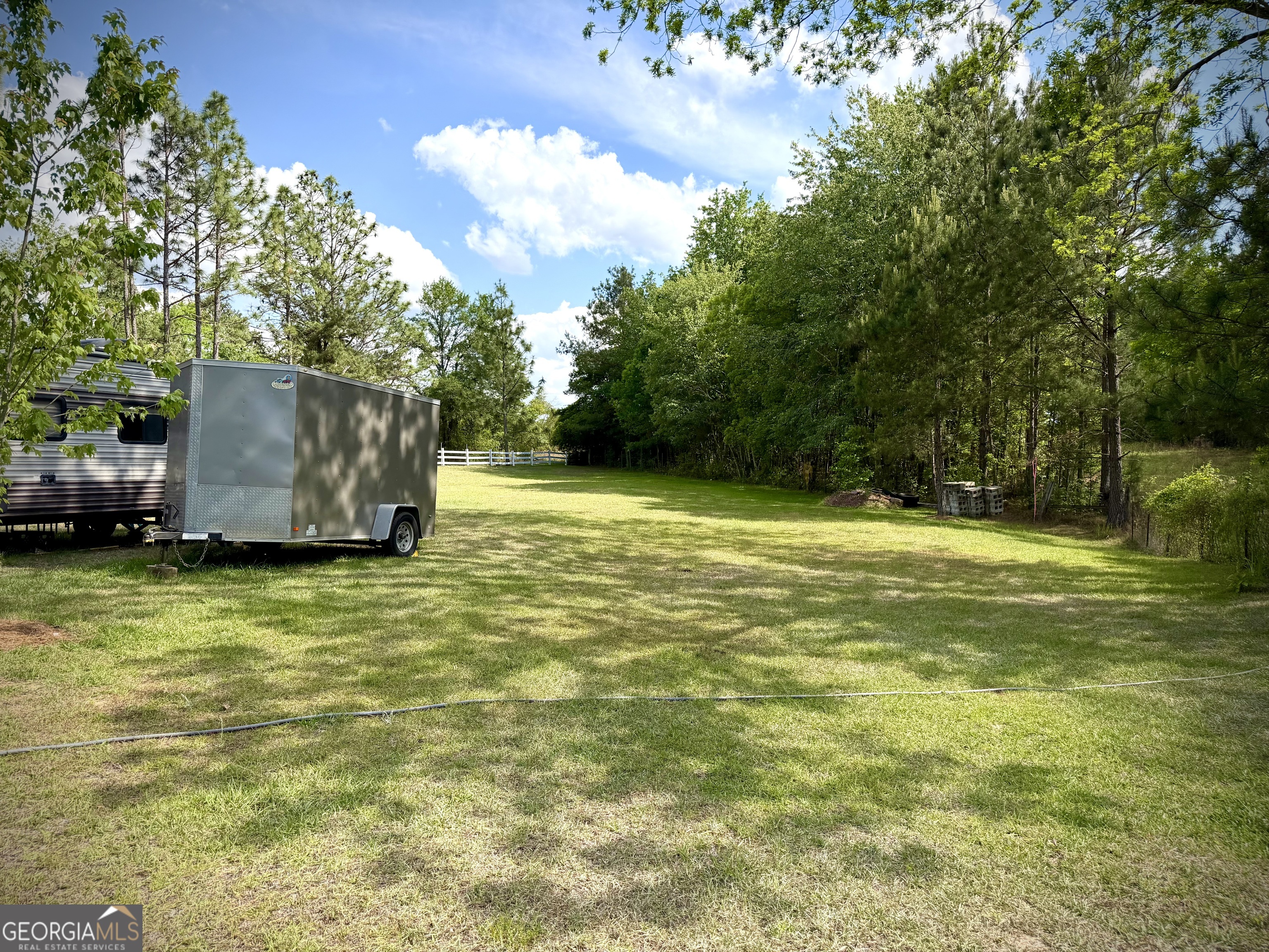 Grassy clearing bordered by trees, with a travel trailer and enclosed utility trailer.