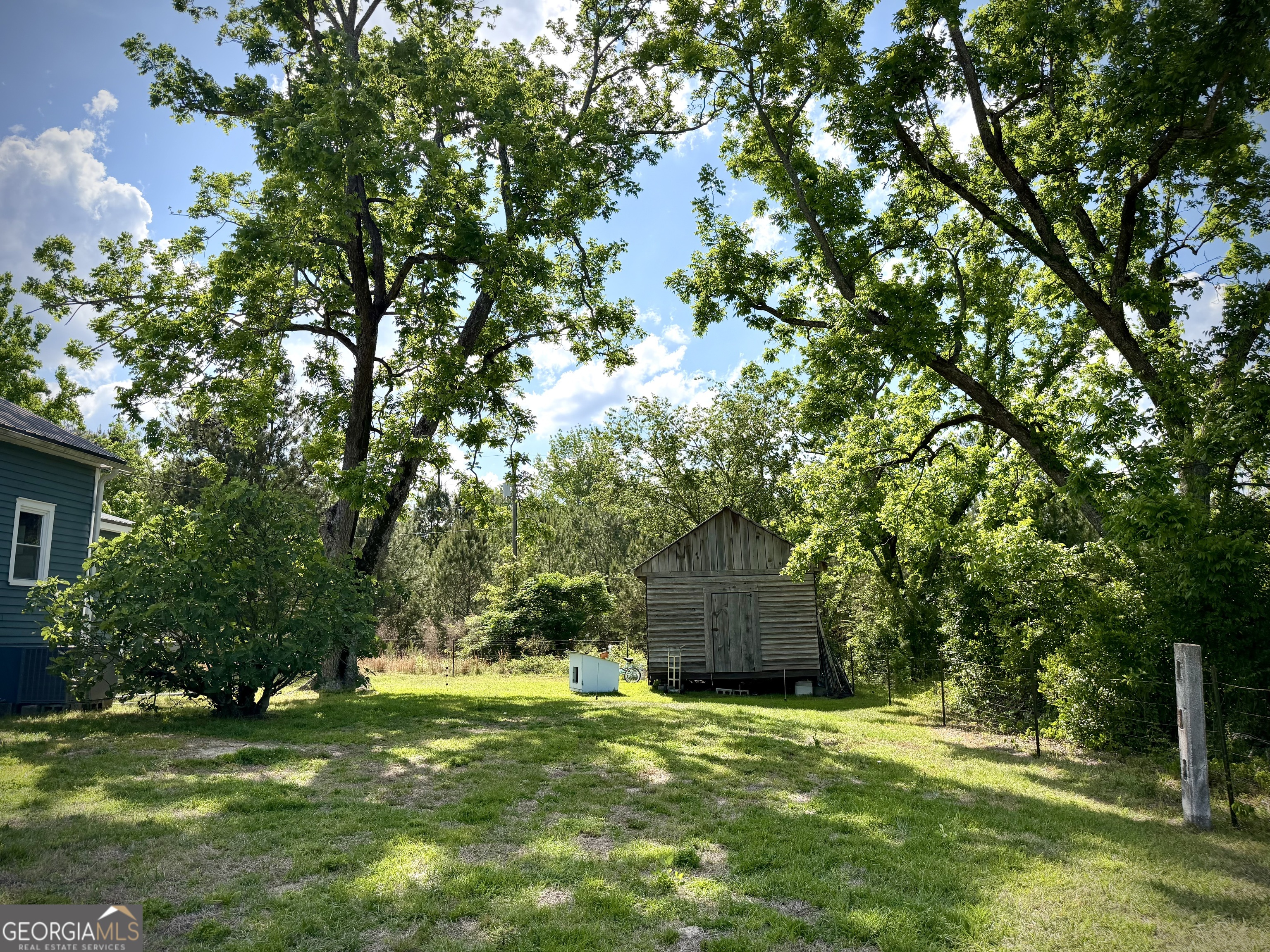 Wooden outbuilding in a grassy, tree-shaded yard beside a blue-sided house.