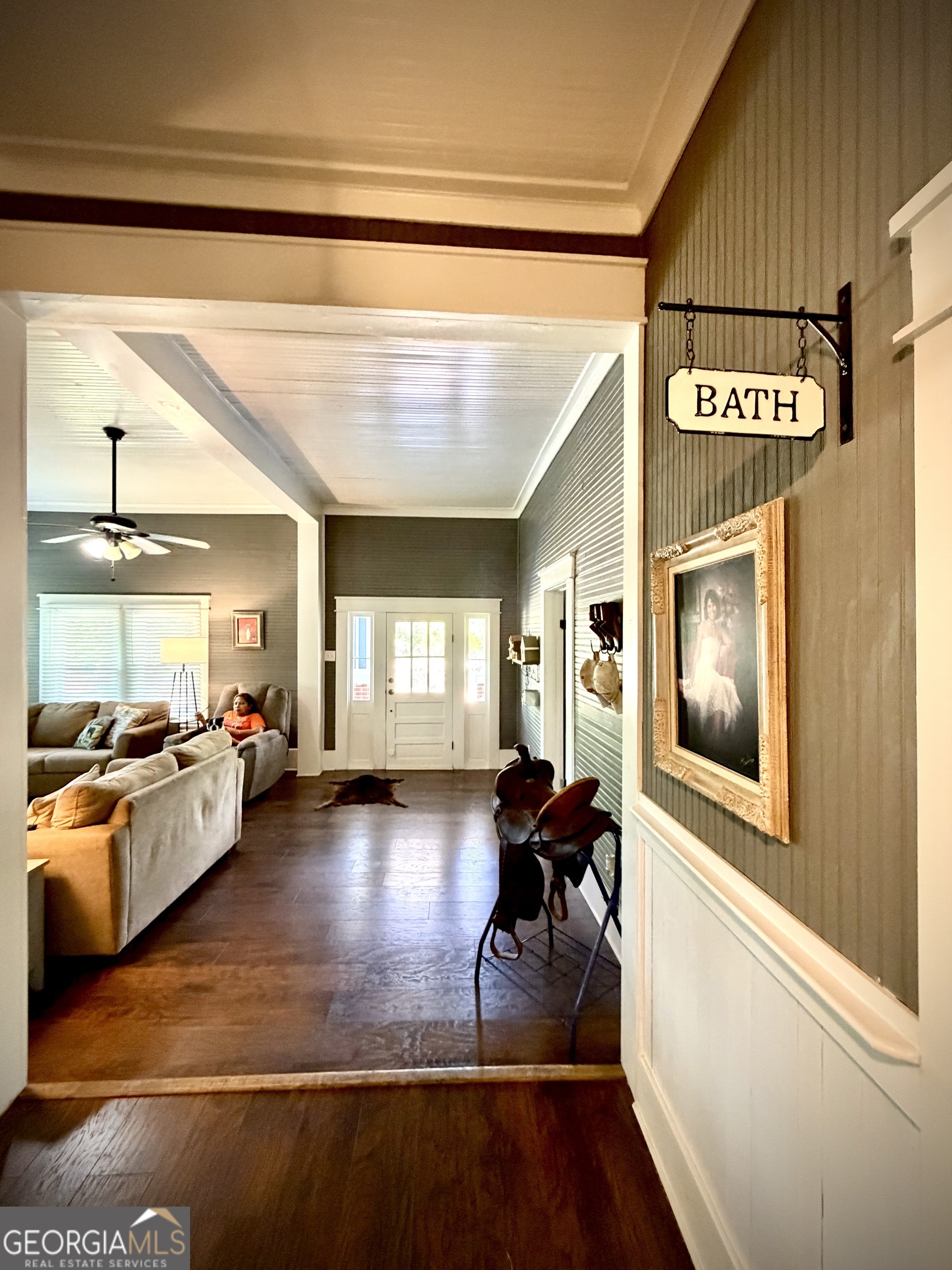 Entry hall opens into a furnished living room, with a hanging bath sign and saddle display.