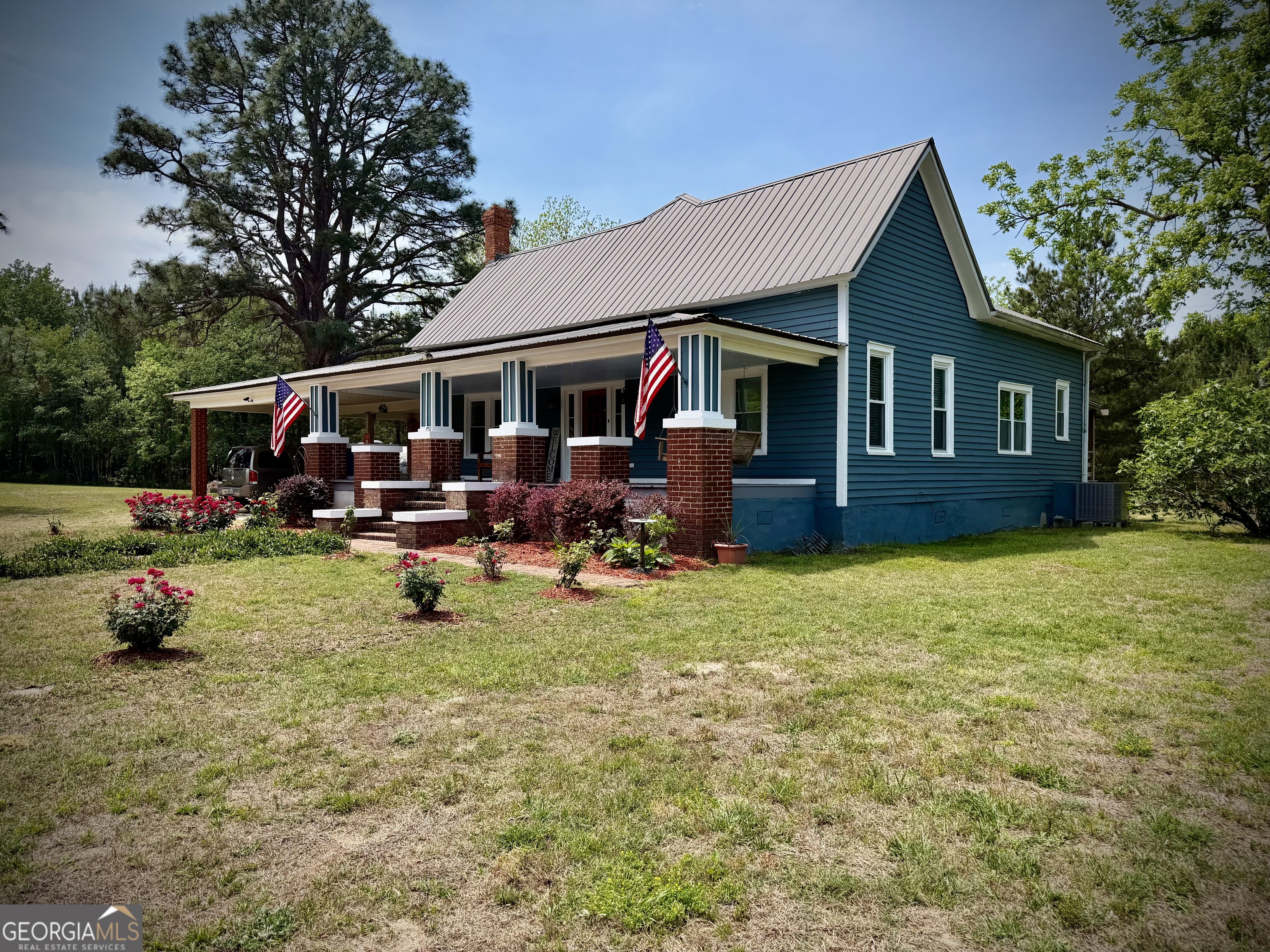 Teal-blue house with a wide brick-columned porch, American flags, and flowering shrubs.