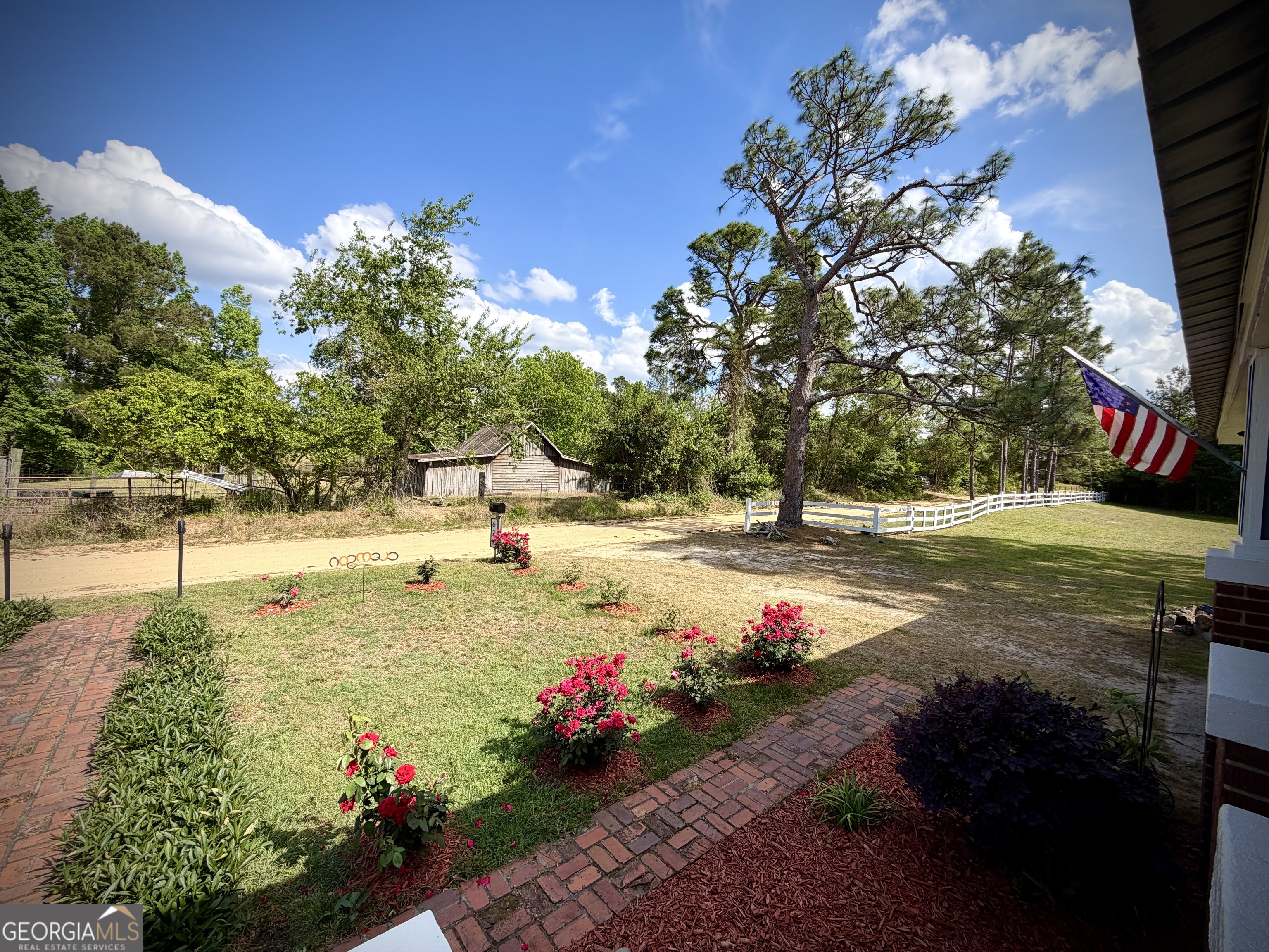 Landscaped yard with brick paths, rose bushes, a large pine tree, white fencing, and an American flag.